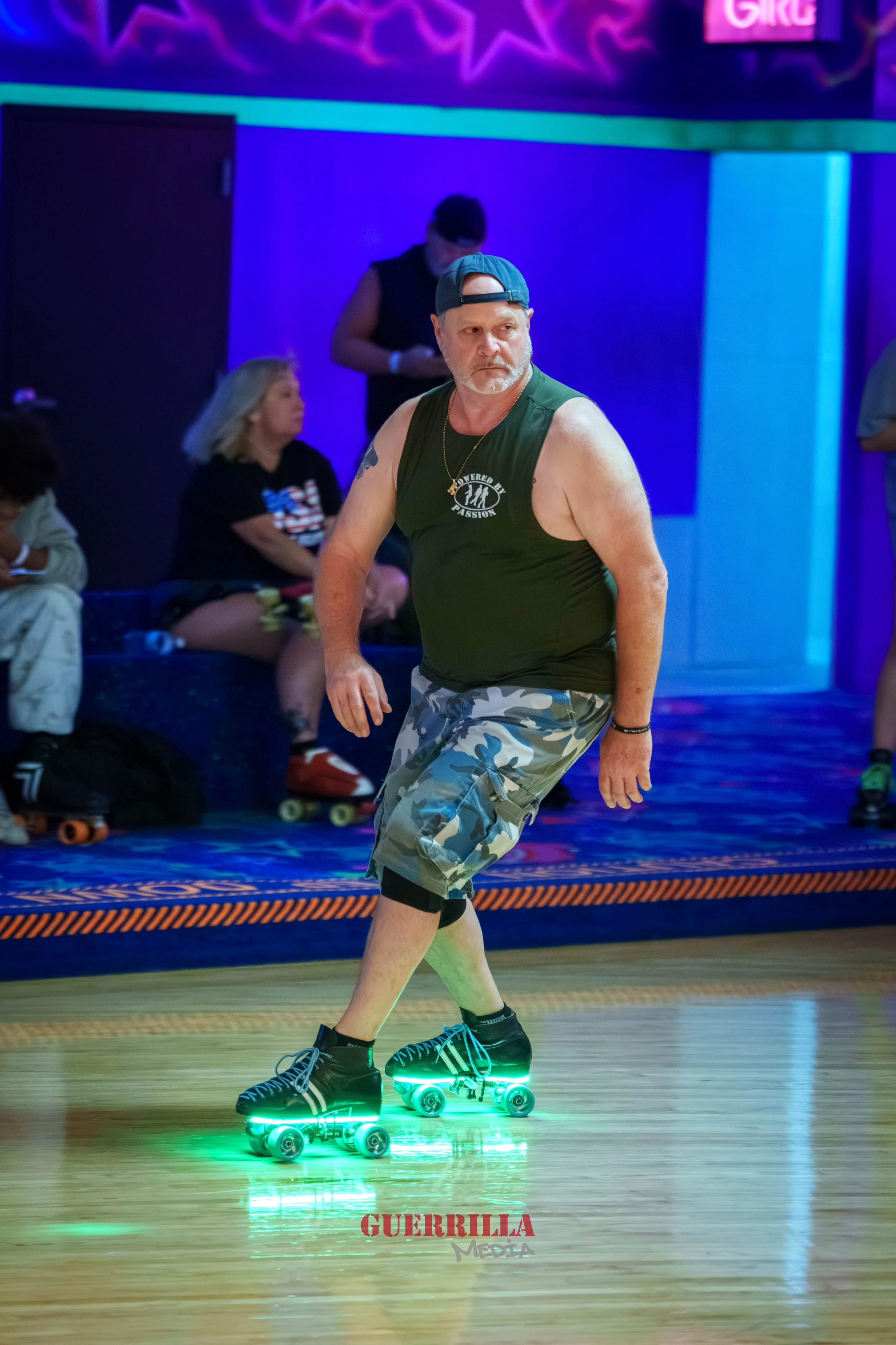 A middle-aged man roller skating at an indoor roller rink, with neon lights and people sitting in the background.