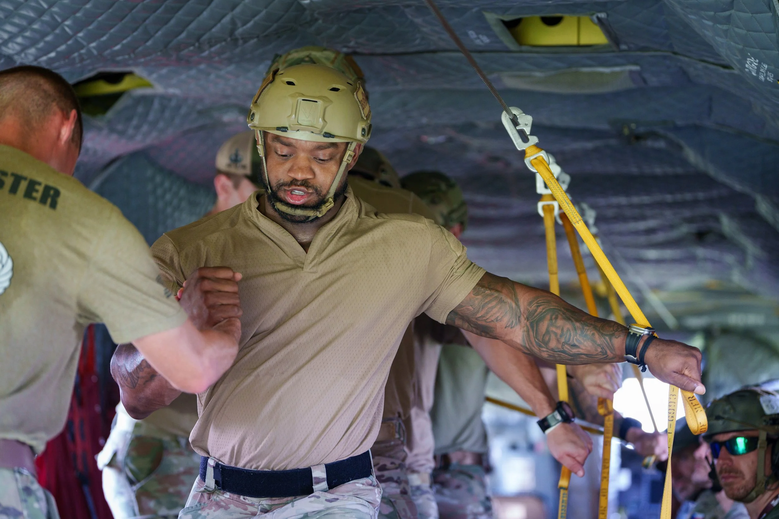 Soldiers wearing tan uniforms and helmets in a military setting under a tarp, assisting each other with a stretch or exercise, with a focus on one soldier on the right holding a measuring tape.