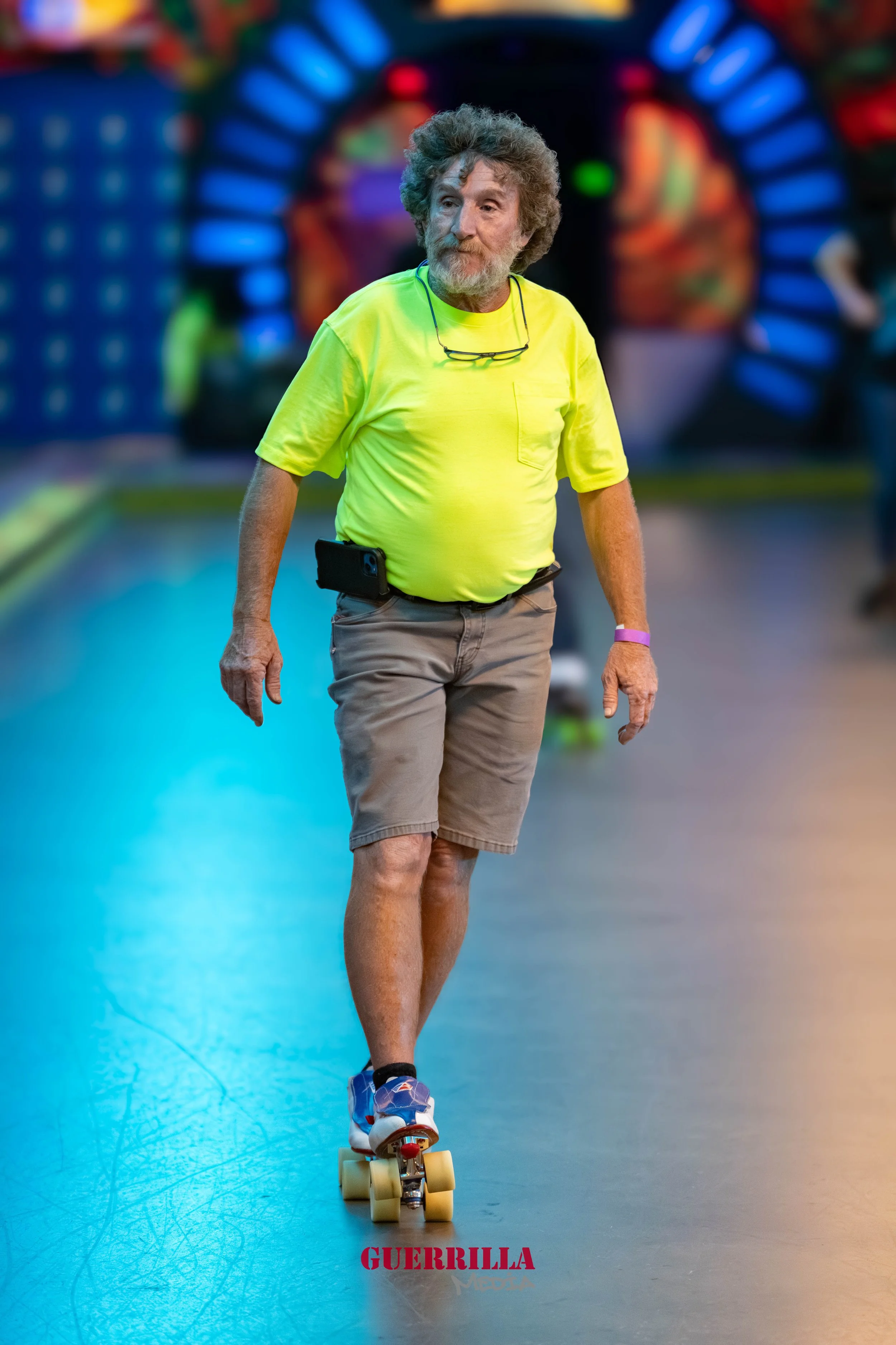 An elderly man with curly gray hair and beard skateboarding indoors, wearing a bright yellow T-shirt, gray shorts, and sneakers, with glasses hanging around his neck.