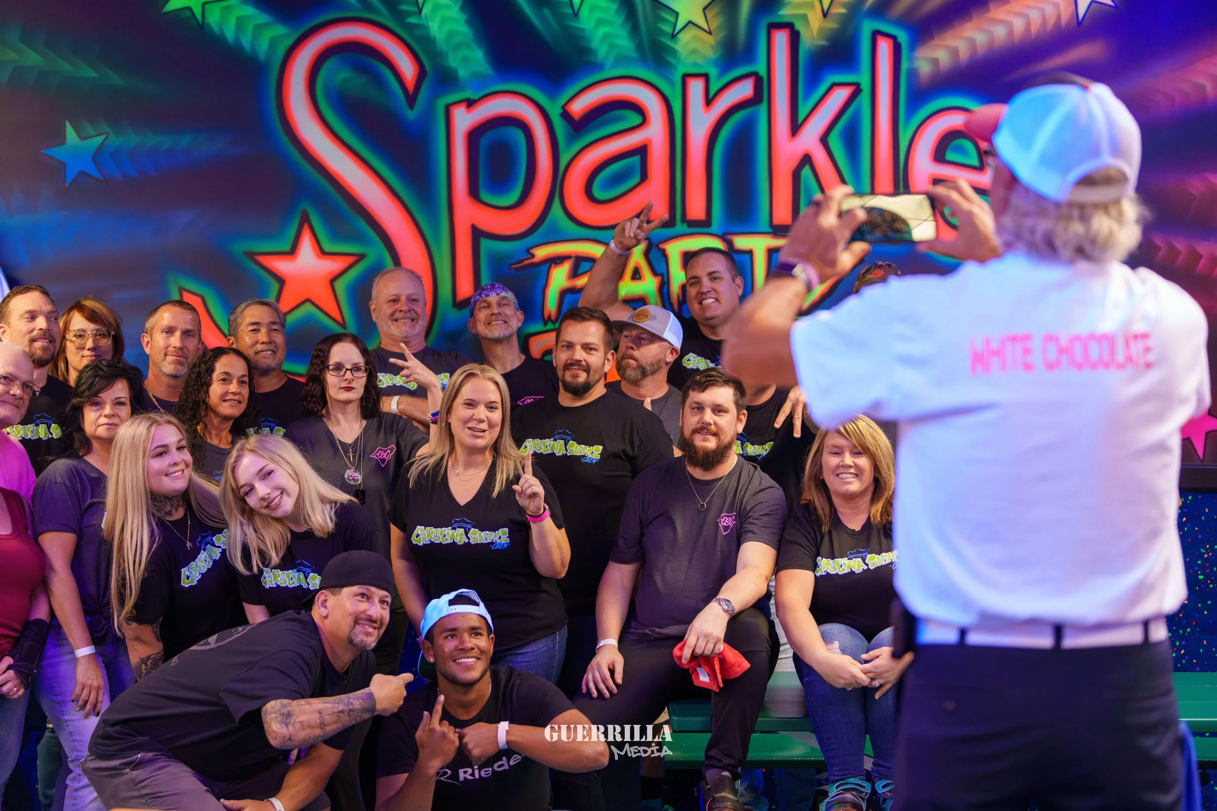 Group of people posing for a photo at an event called Sparkle Party, with a person taking a picture in the foreground wearing a white shirt that says 'White Chocolate', and a colorful backdrop featuring the event's name.