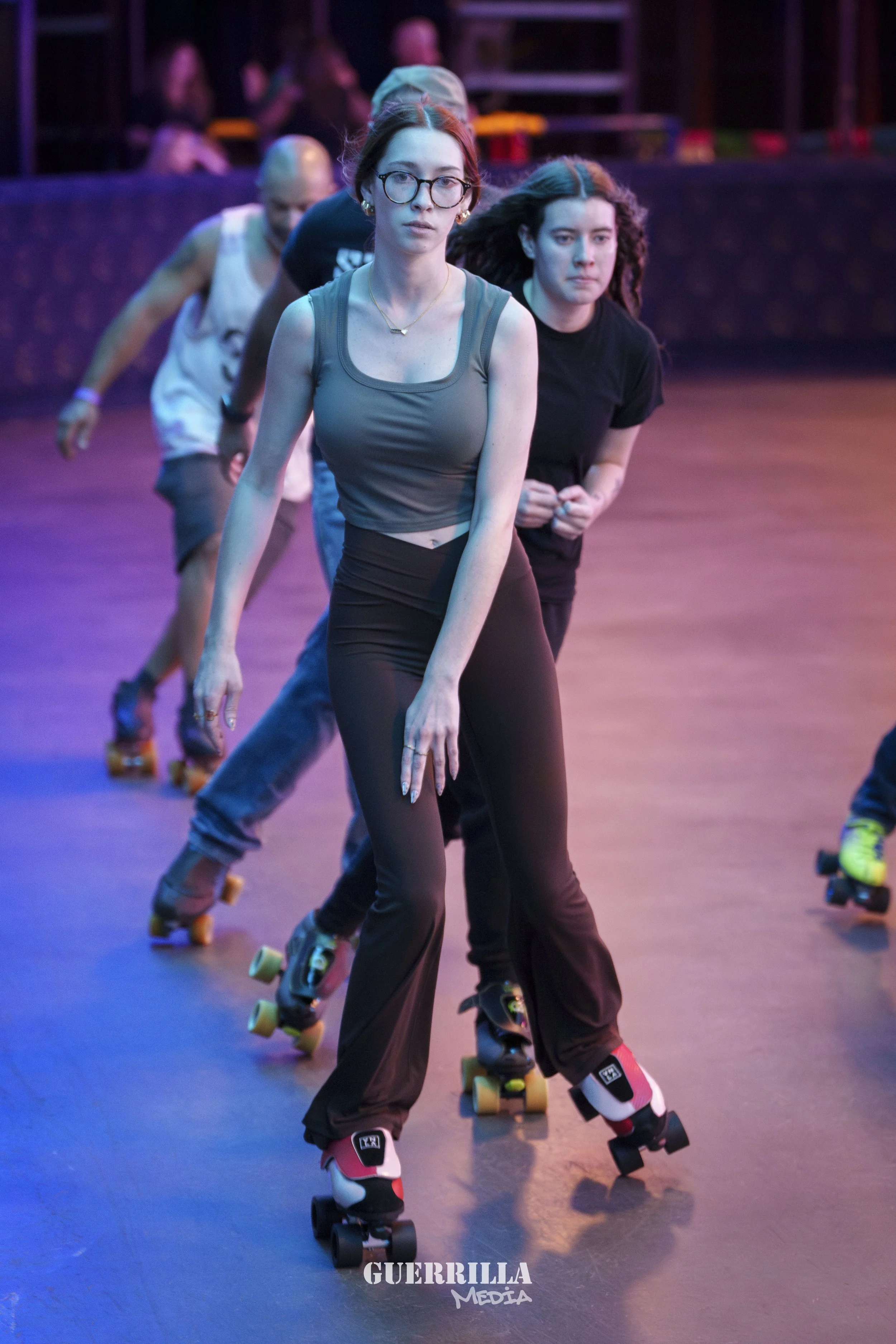 Group of people roller skating indoors under colorful lighting, with a focus on a young woman in the front wearing glasses, a gray tank top, and black pants.