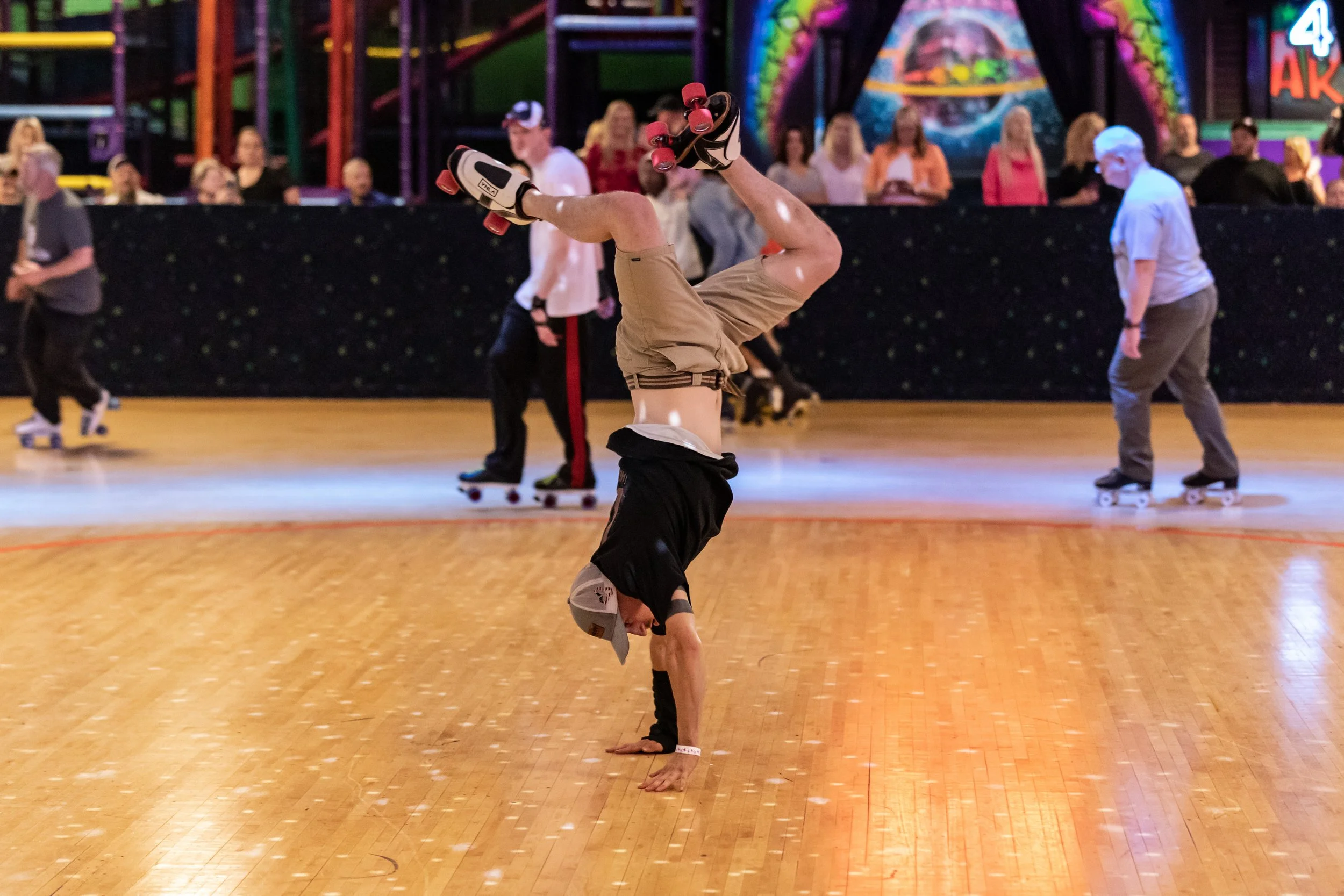 A person practicing breakdancing in a roller skating rink, balancing on one hand with legs in the air, wearing a helmet and knee pads, with other skaters and spectators in the background.