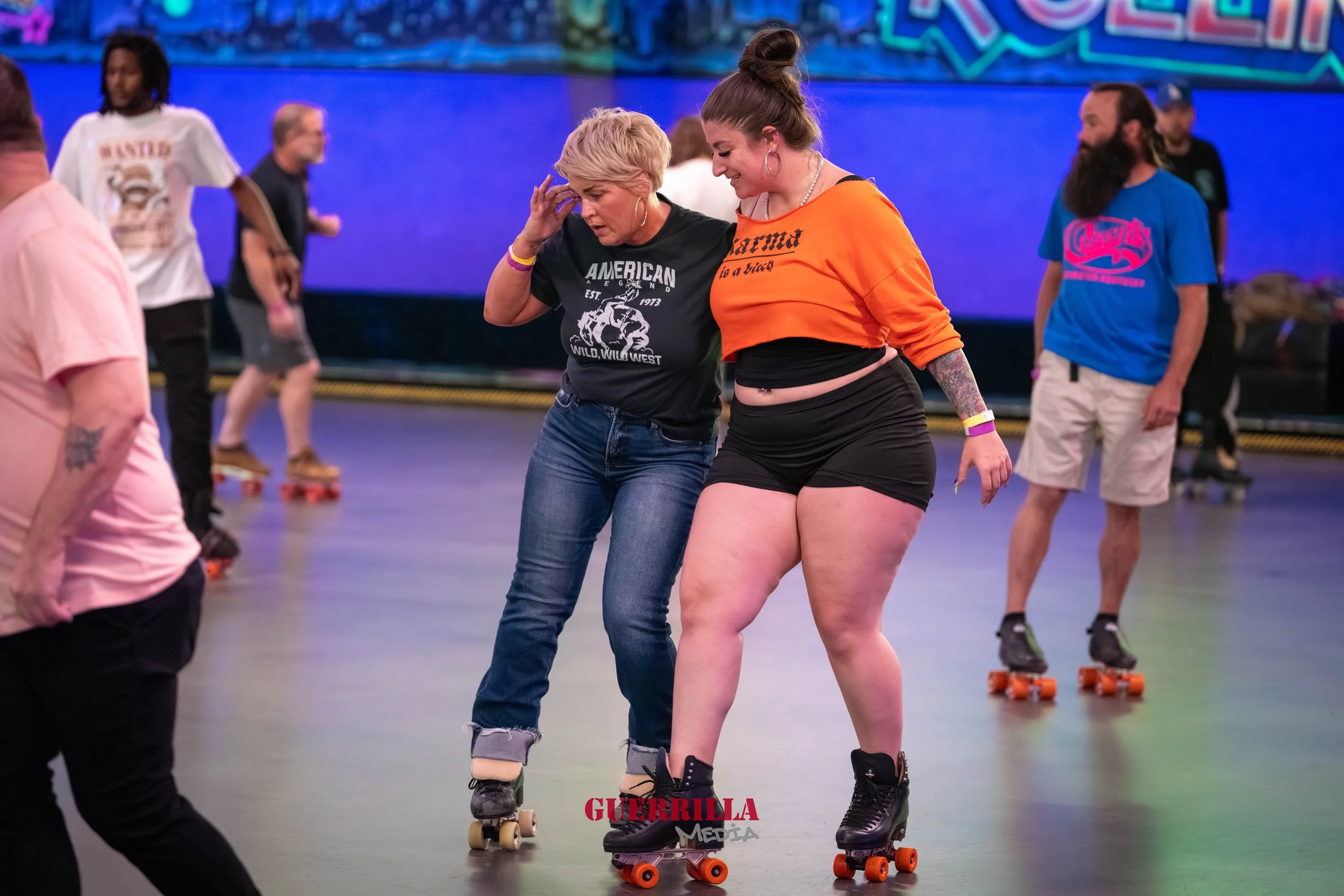 A woman in an orange crop top and black shorts rollerskates with a woman in a black t-shirt and jeans, both smiling and enjoying roller skating in an indoor rink, with other skaters in the background.