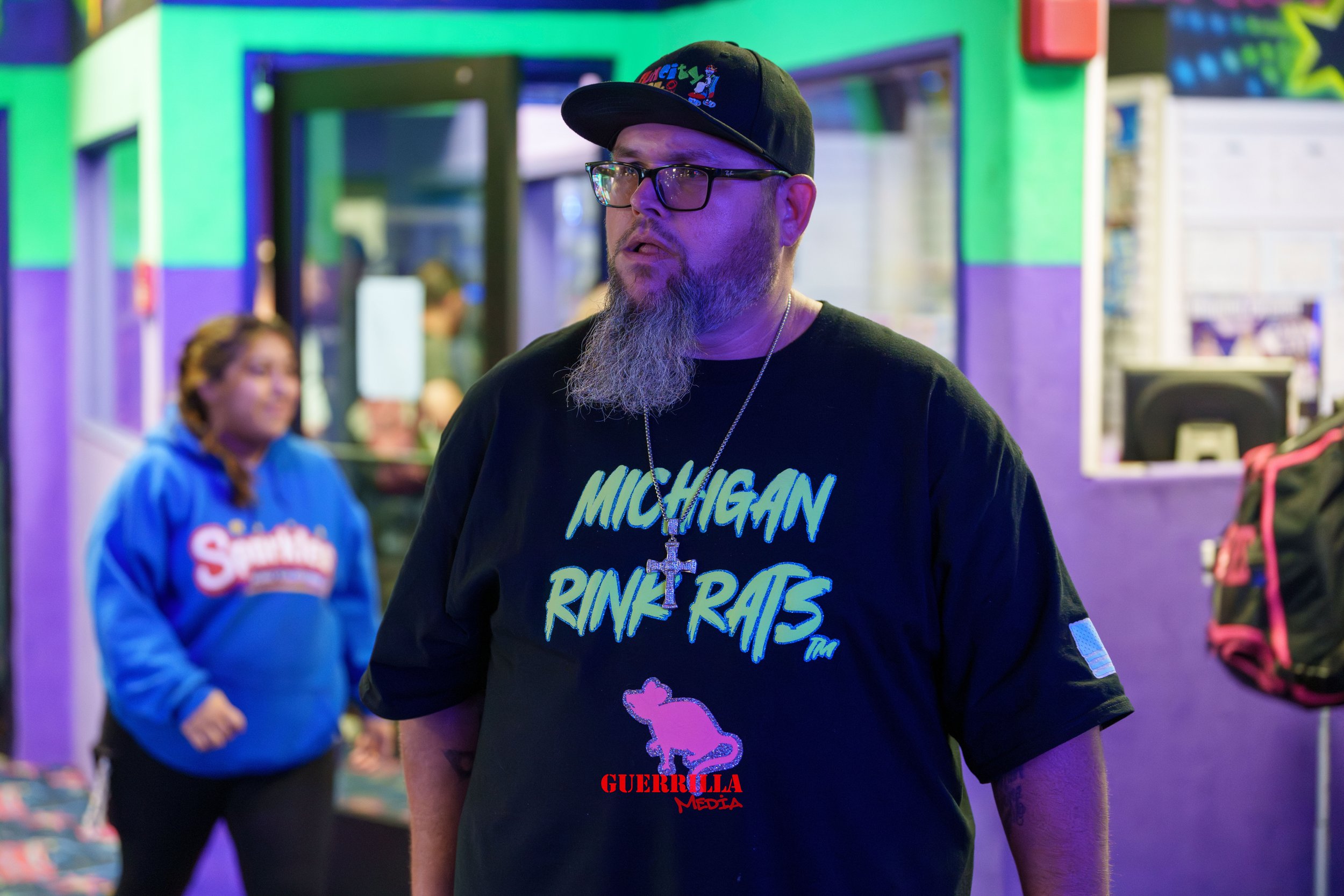 A man with glasses and a beard wearing a black cap and a black T-shirt with the words "Michigan Ringers" in neon style font and a pink pig graphic. He has a silver cross necklace and appears to be inside a colorful arcade or entertainment center. In 