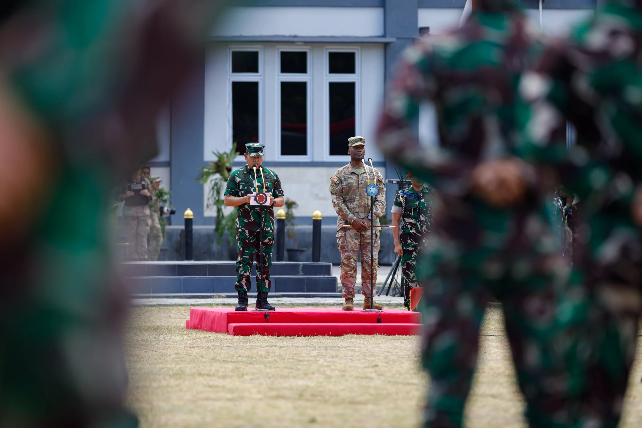 Military officials and soldiers standing on a platform during a formal outdoor ceremony, with others in formation visible on the sides.