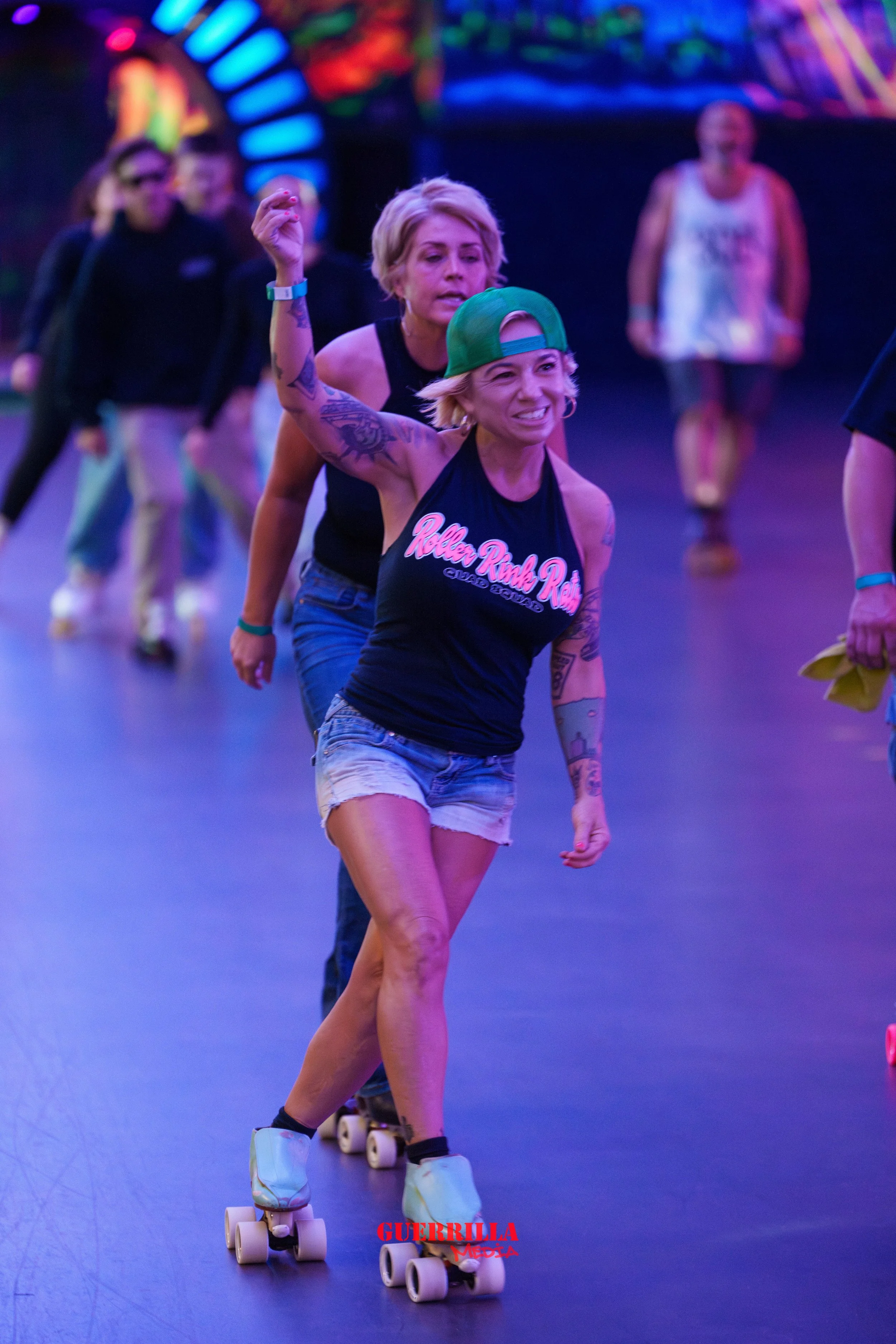 A group of people roller skating indoors under colorful neon lights. The woman at the front is smiling, wearing a green cap backward, a black tank top, and denim shorts.