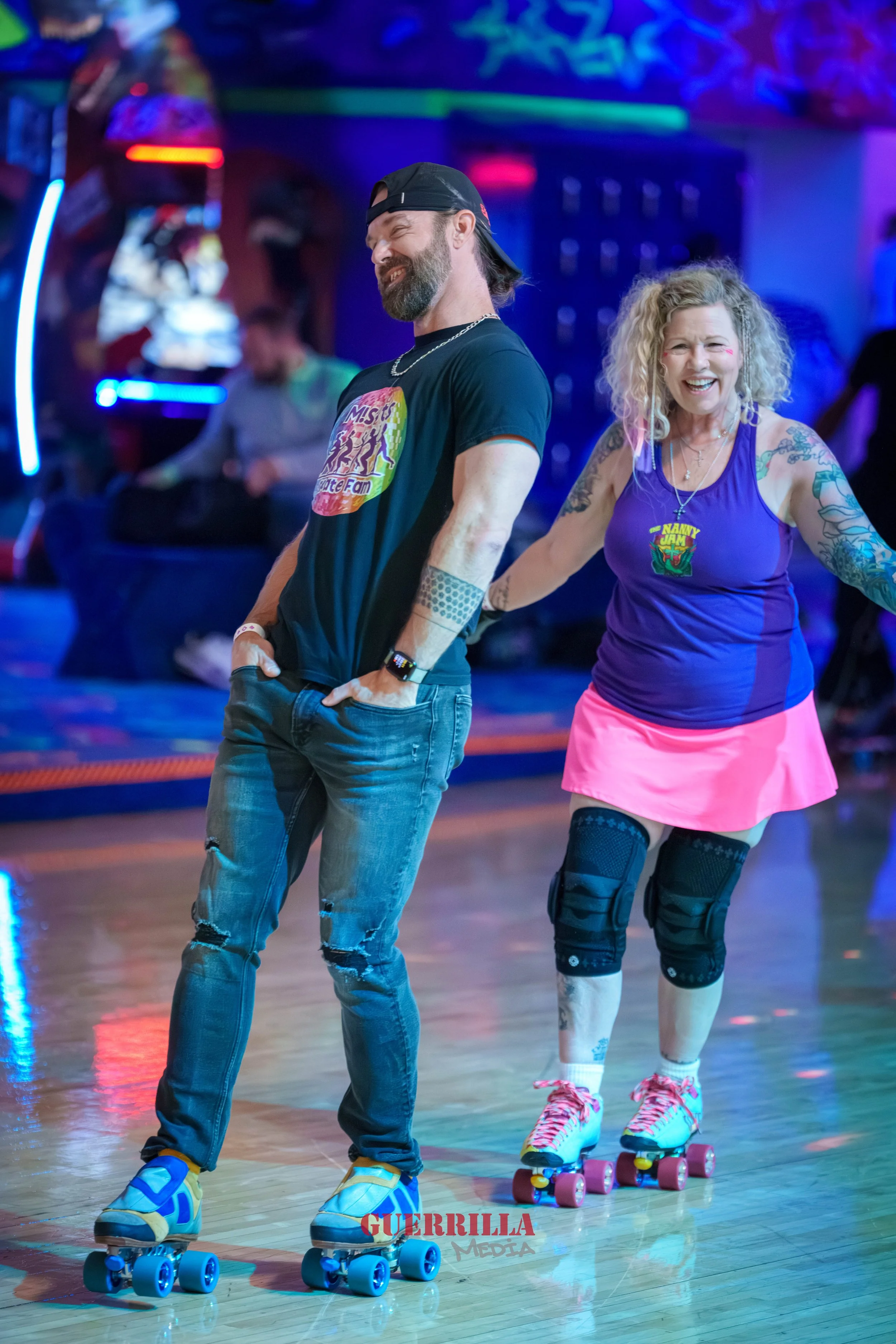 Two people roller skating in a colorful, vibrant indoor roller rink, smiling and enjoying themselves. The man has a beard, is wearing a black cap backwards, a black t-shirt, ripped jeans, and blue roller skates. The woman has curly gray hair, tattoos