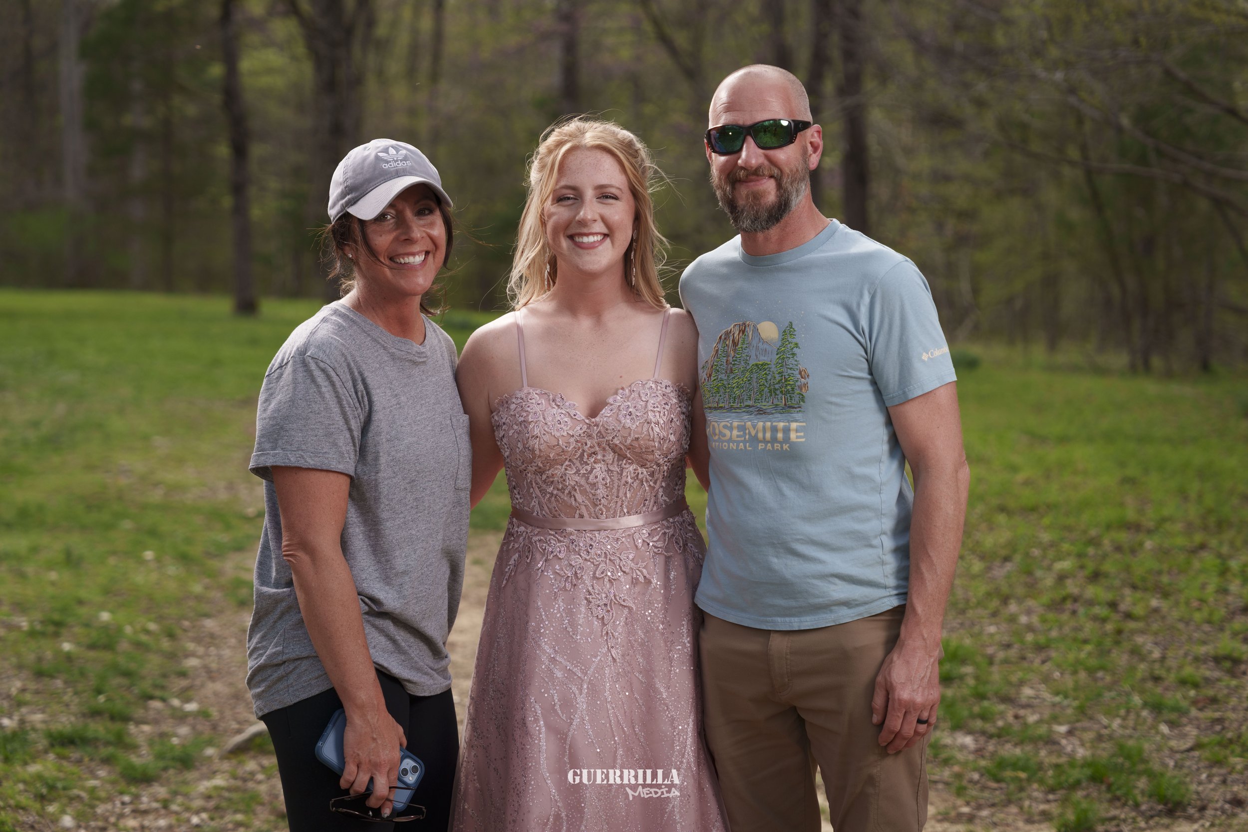 Three people standing outdoors in a wooded area, smiling at the camera, with a woman in a pink lace dress in the center, a woman in a gray t-shirt and baseball cap on the left, and a man in a blue t-shirt with a "Yosemite National Park" graphic and s