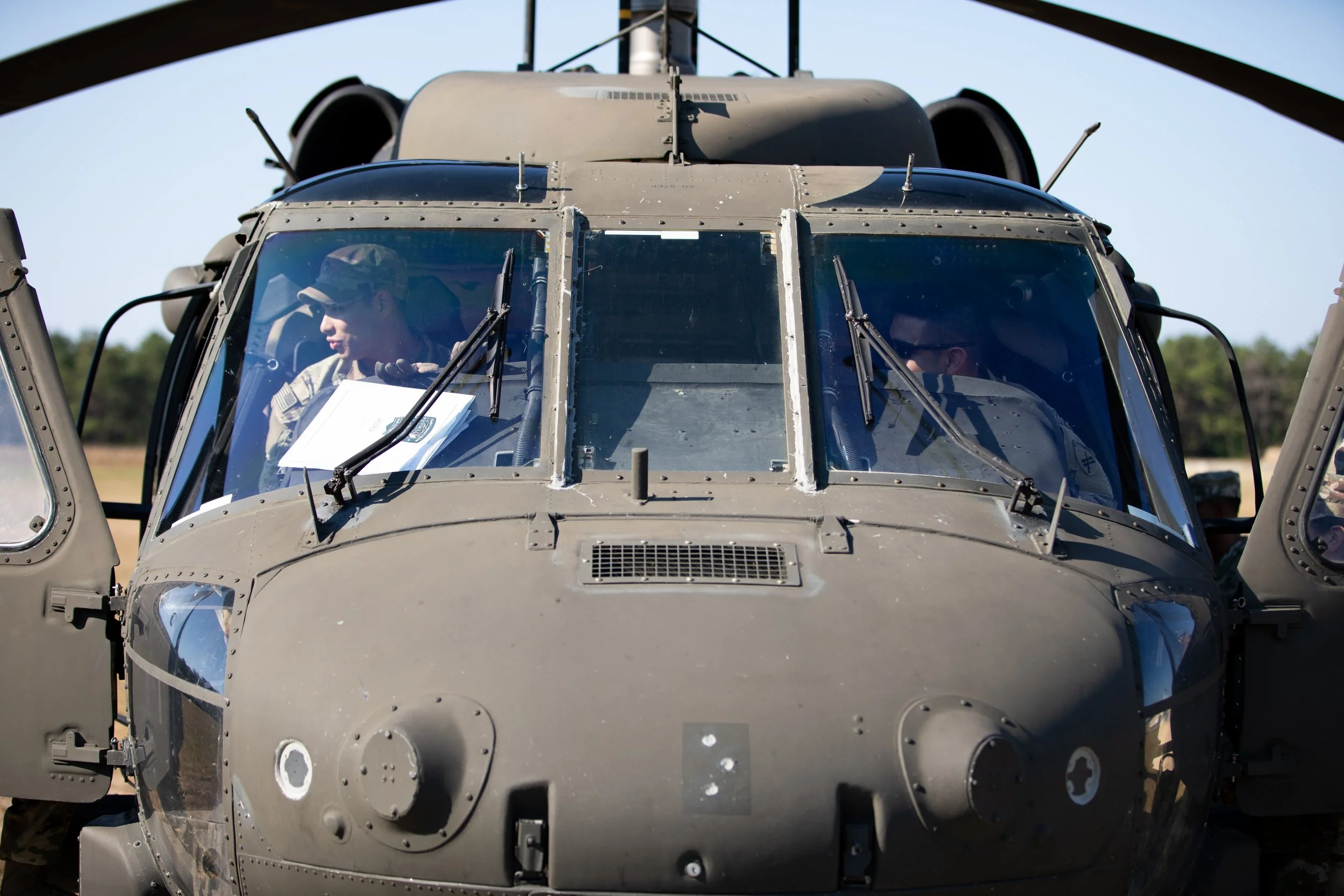 Front view of a military helicopter with two soldiers inside, one reading a document, on a clear day with trees in the background.