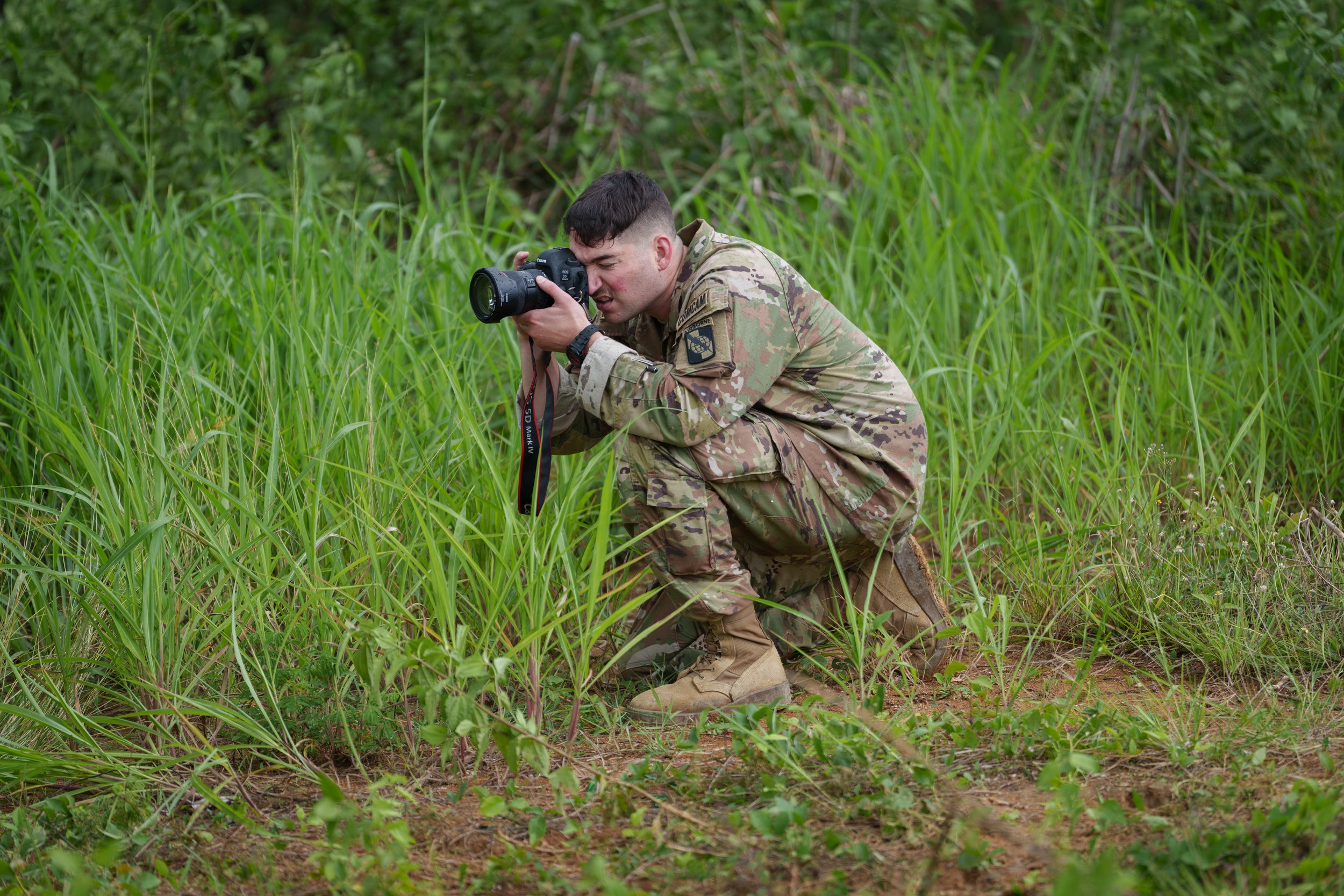 A soldier in camouflage uniform kneeling in tall green grass, taking a photograph with a camera.