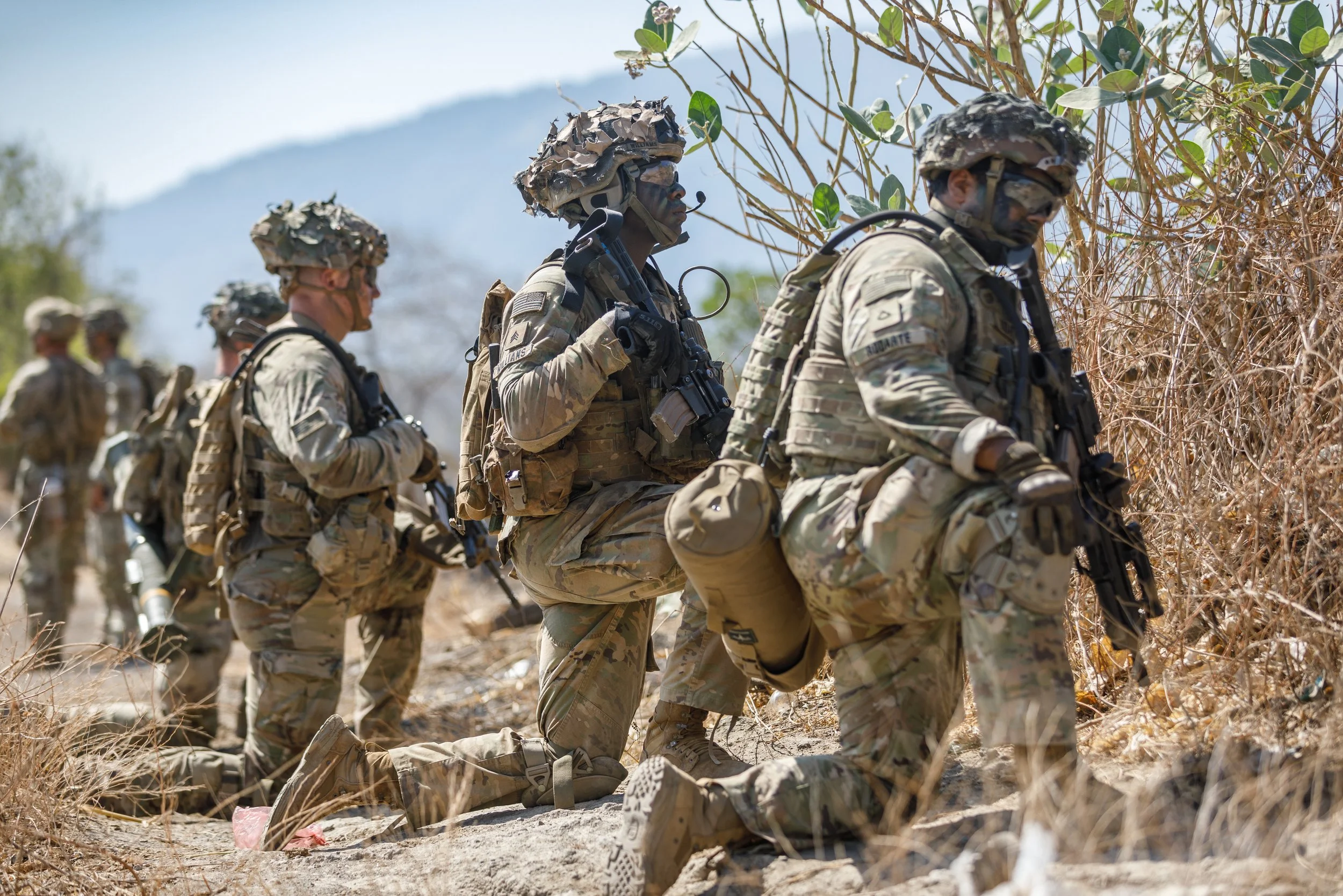 Military soldiers in camouflage uniforms kneeling in a line during a training exercise in a dry, outdoor environment with mountains in the background, holding rifles.