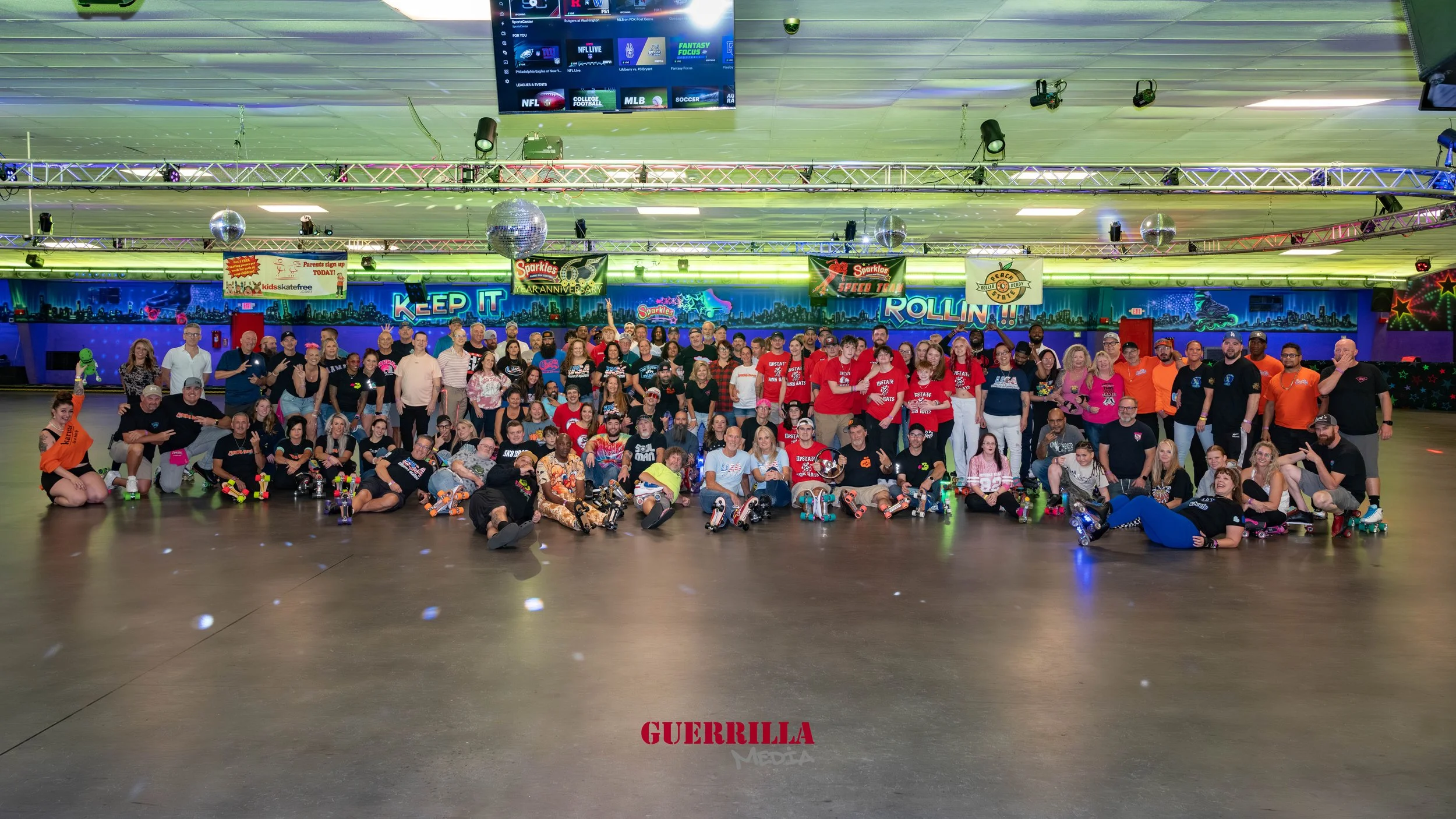 A large group of people gathered at a roller skating rink for a group photo, with some wearing matching team shirts and roller skates on the floor in front of them. The background features colorful neon signs and a digital screen displaying sports an