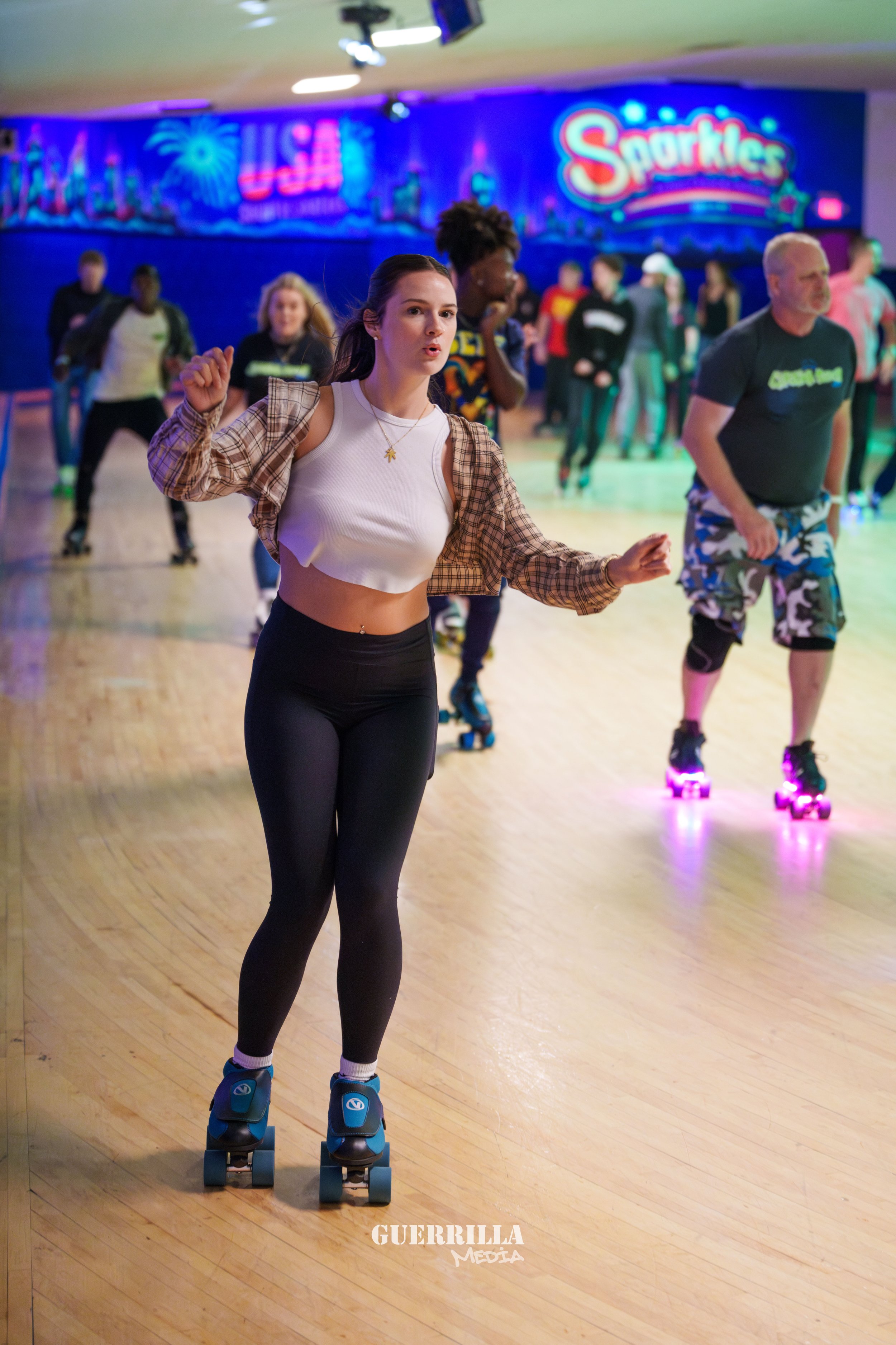 A young woman roller skating at an indoor roller rink, wearing a white crop top and black leggings, with a brown checked shirt over her shoulders and a necklace with a star pendant. The rink has neon signs in the background, including one that says '