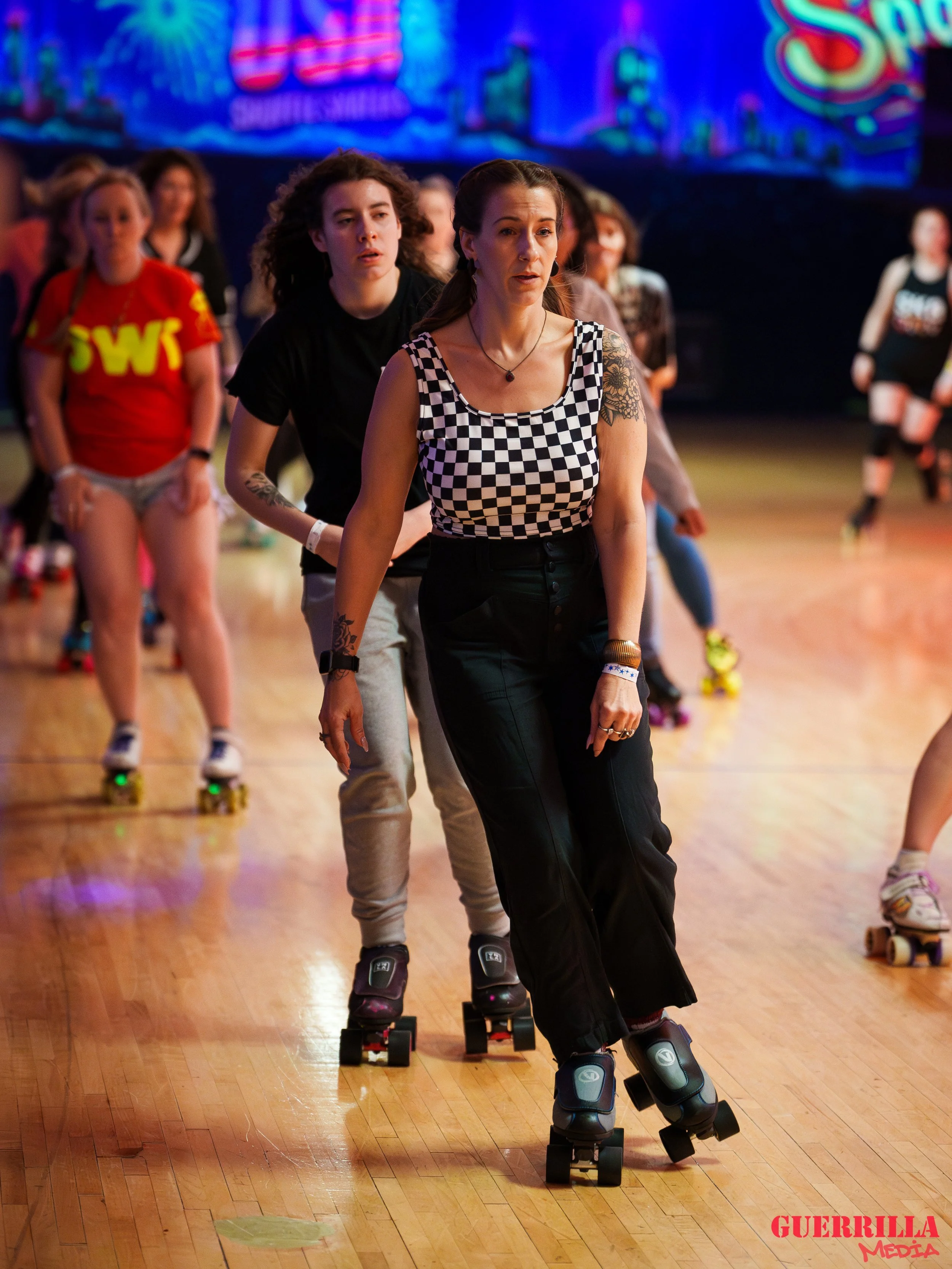 People roller skating at an indoor rink, with a woman leading in the foreground and others following behind. The background has colorful lights and a vibrant sign.