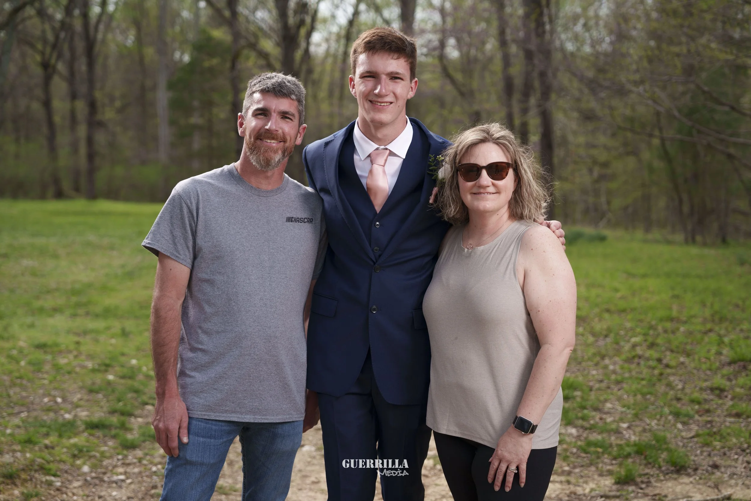 A family of three standing outdoors on a grassy area with trees in the background. The man on the left has gray hair, a beard, and is wearing a gray t-shirt. The young man in the middle is dressed in a suit with a pink tie. The woman on the right is 