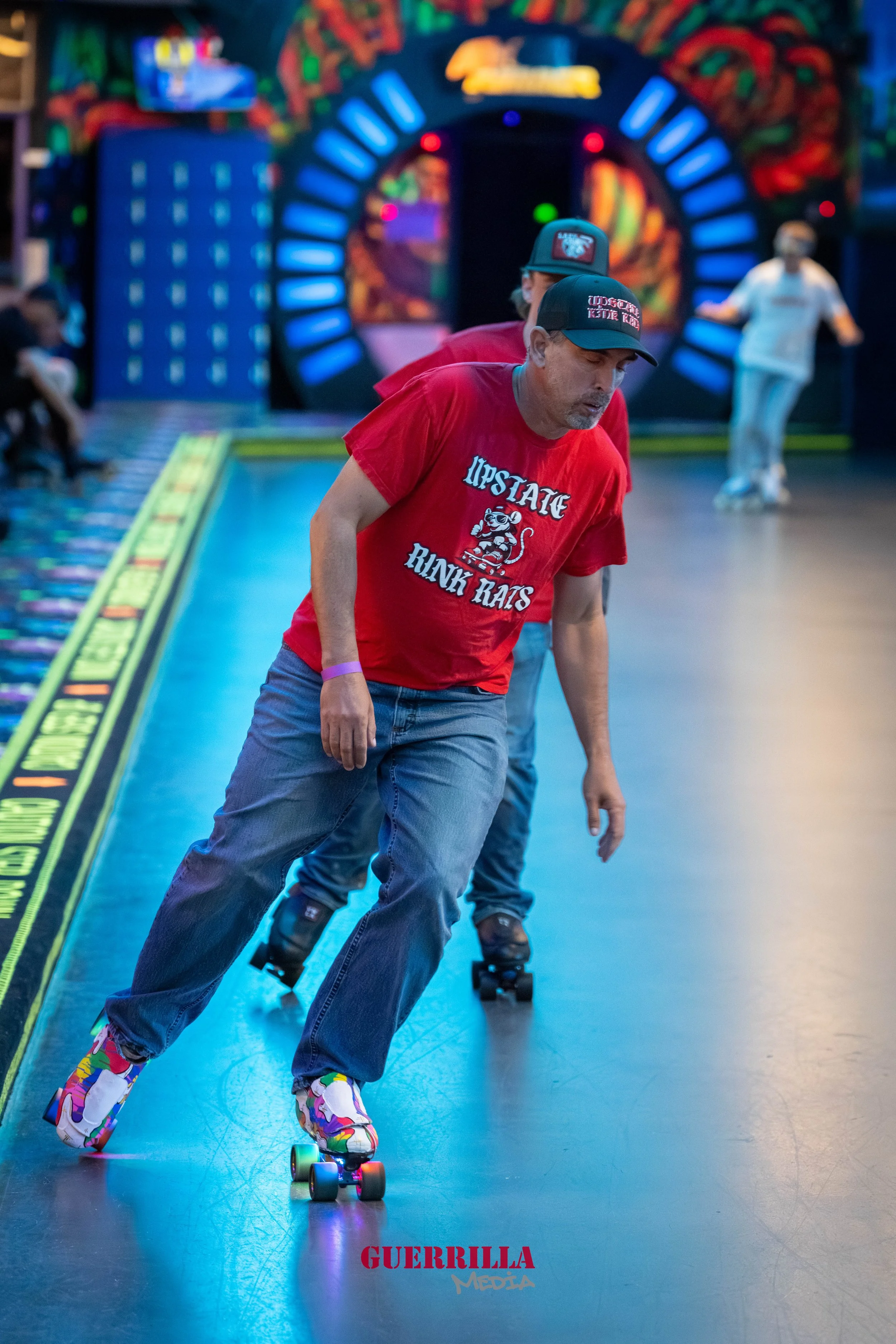 Two men roller skating indoors, with a colorful, vibrant background featuring a neon-lit arcade and abstract wall art. The man in front is wearing a red T-shirt, gray jeans, and colorful roller skates.