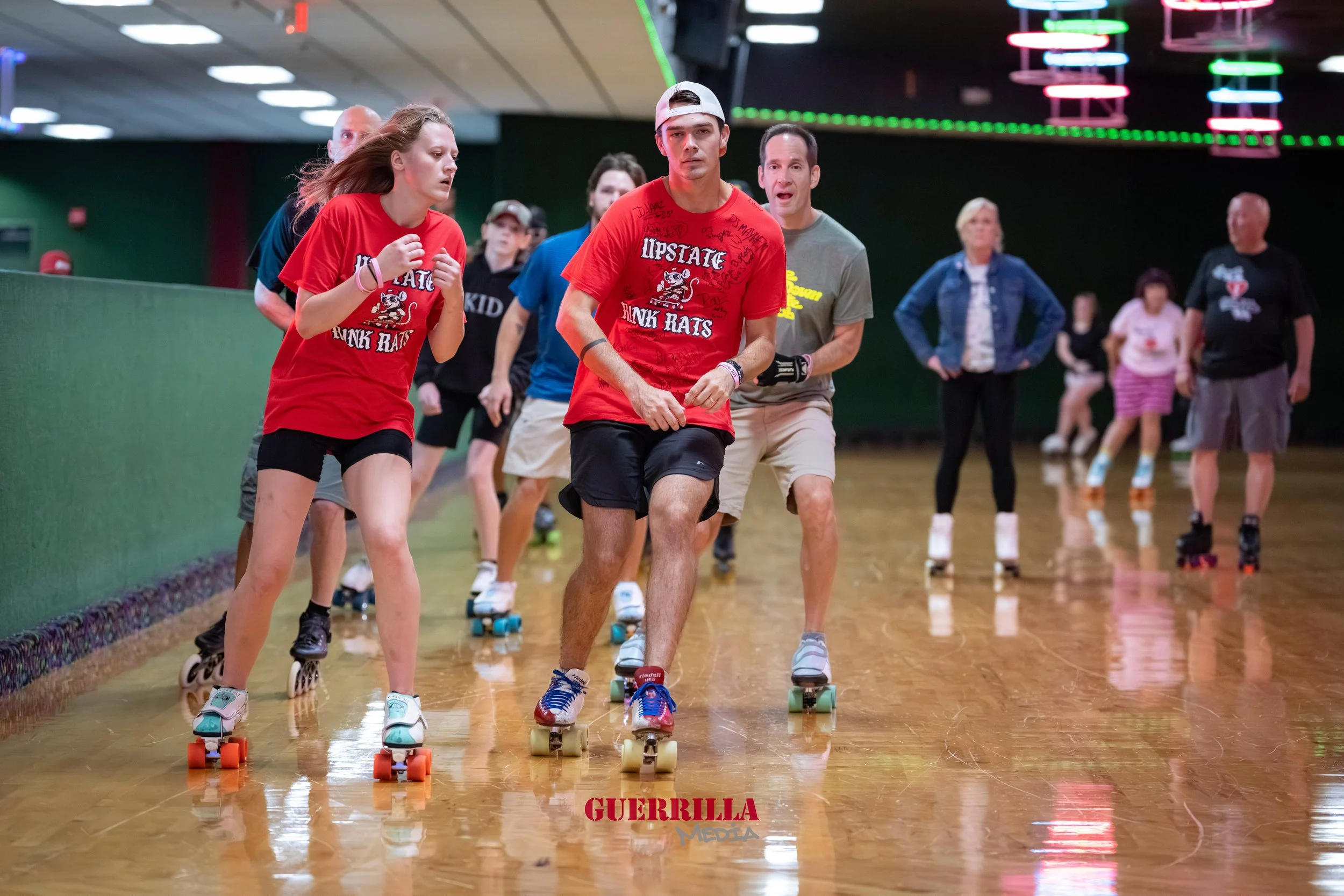 People roller skating indoors, with a young man in the foreground wearing a red t-shirt, black shorts, and a backward cap, leading a group of skaters. The skaters in the background include men and women, some watching and some skating.