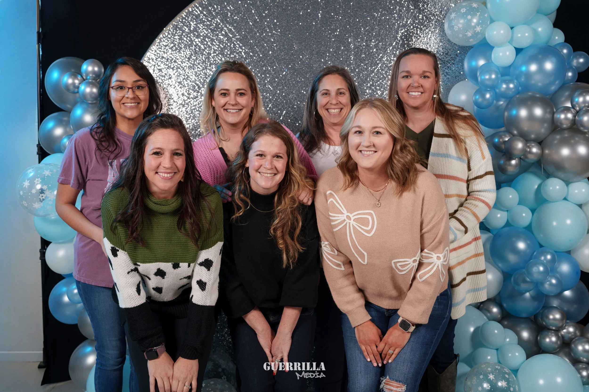 Eight women smiling in front of a festive backdrop with blue and silver balloons, some with snowflake designs, at an event for Guerrilla Media.
