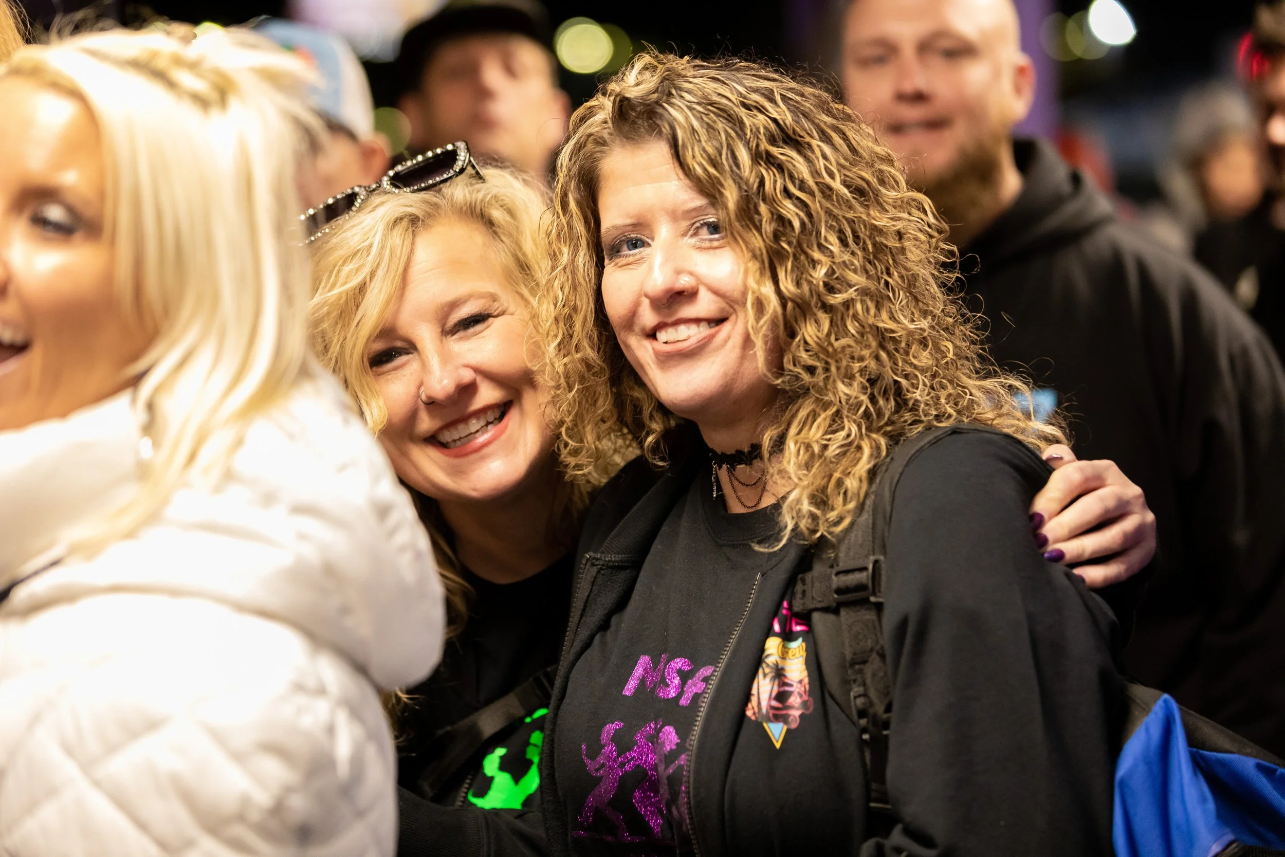 Two women smiling and embracing at a crowded outdoor event during the evening.