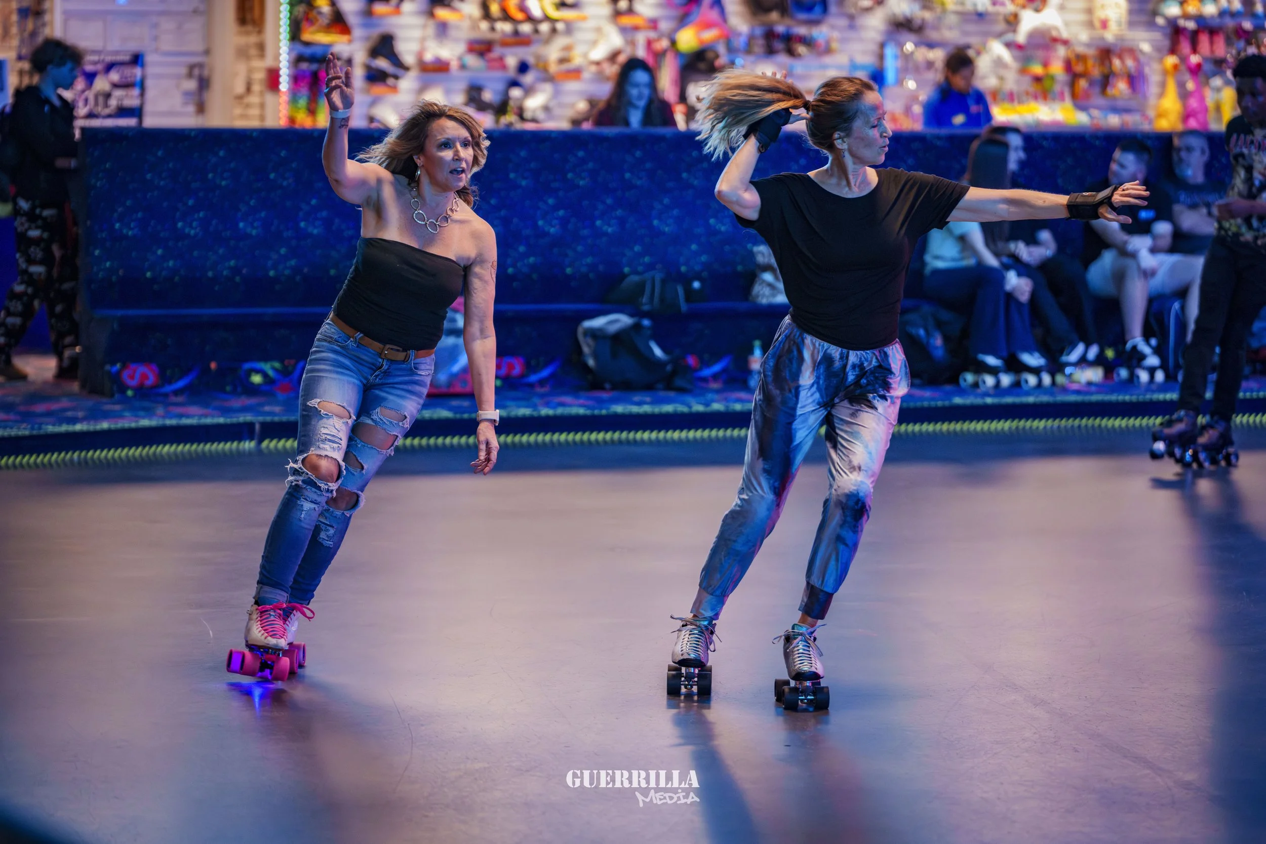 Two women roller skating indoors with a colorful background of arcade games and people, one with curly blonde hair in ripped jeans and a black strapless top, and the other with straight hair in a black t-shirt and patterned pants.