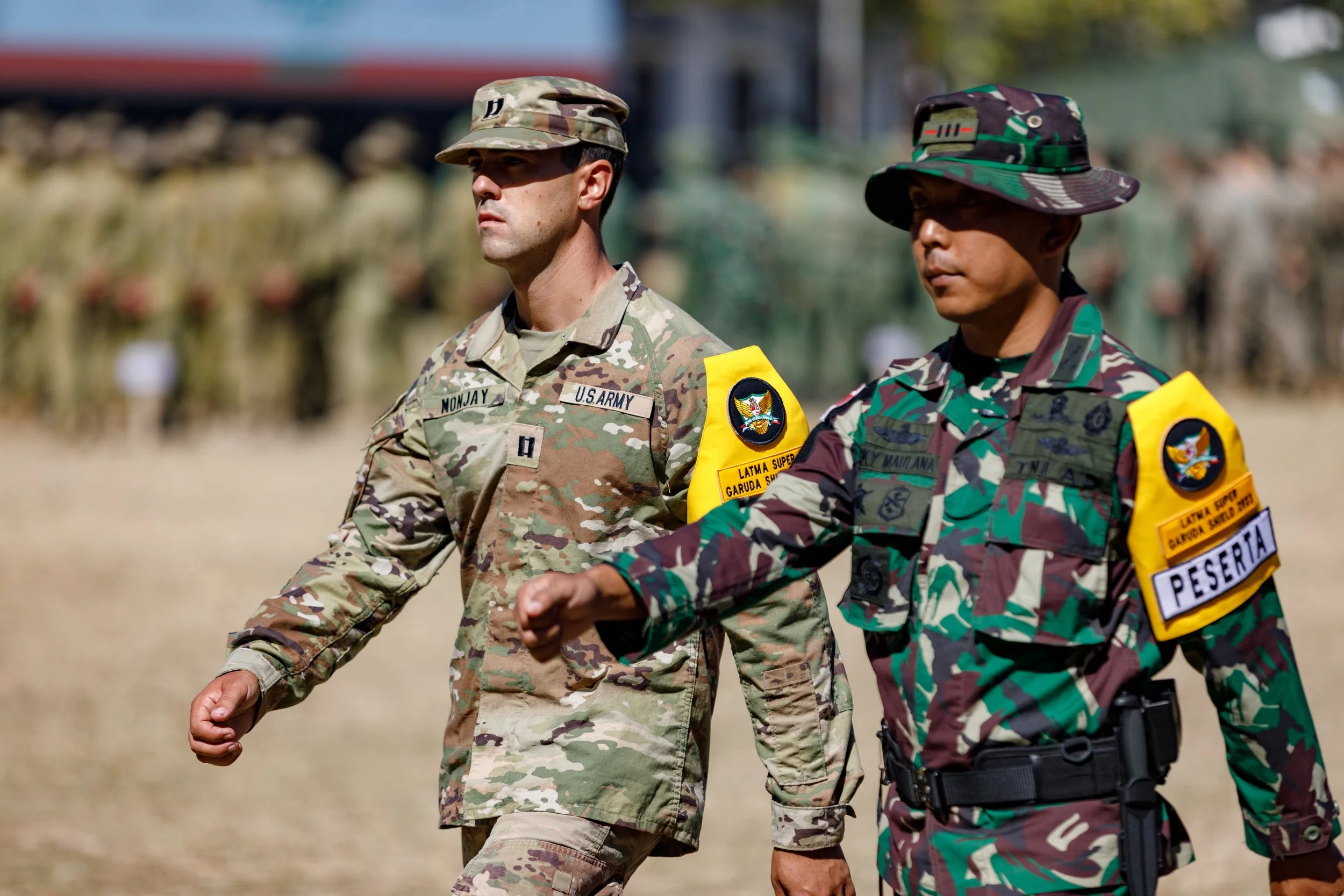 Two soldiers walking outdoors in uniform, one in U.S. Army camouflage and the other in camouflage with a yellow armband labeled 'PESERTA'.