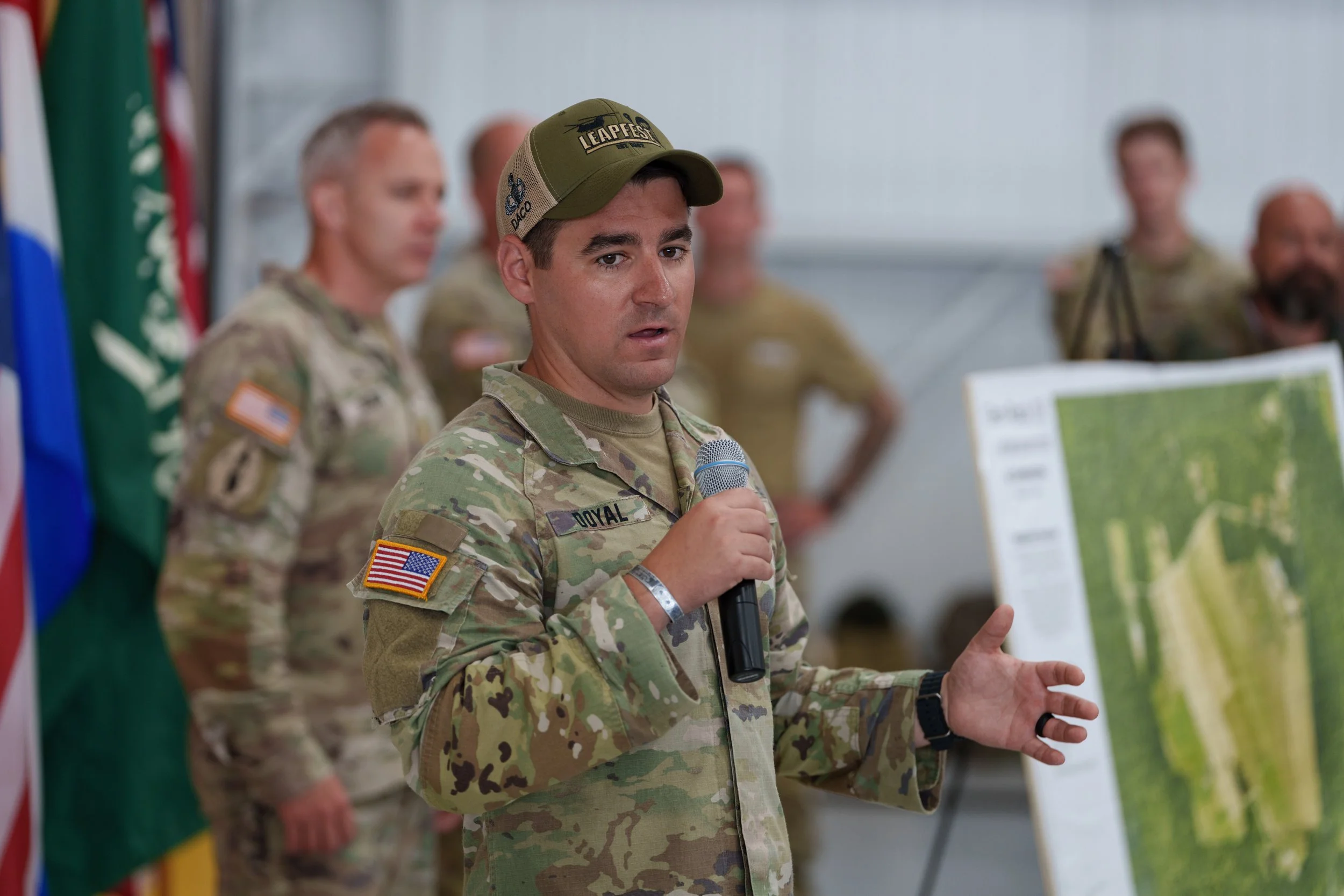 A soldier in camouflage uniform holding a microphone, giving a presentation with a map in front of him, while other soldiers stand behind, with flags visible on the side.