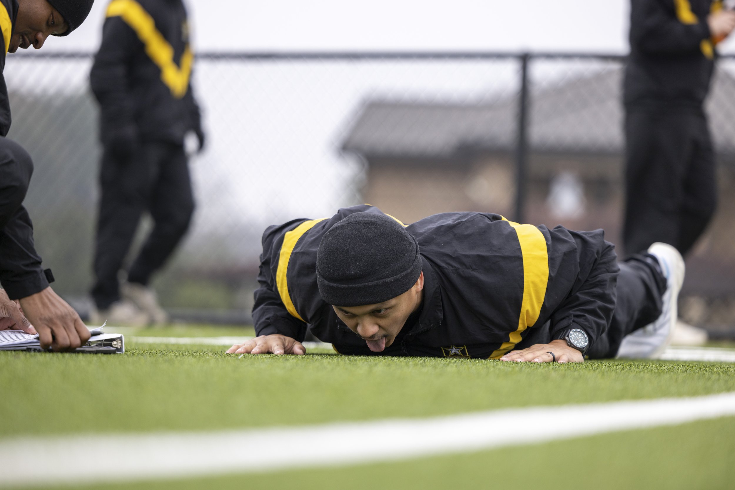 A man in dark athletic clothing with yellow stripes doing a push-up on a green field while sticking his tongue out and wearing a black beanie. Other people in similar athletic clothing are in the background.