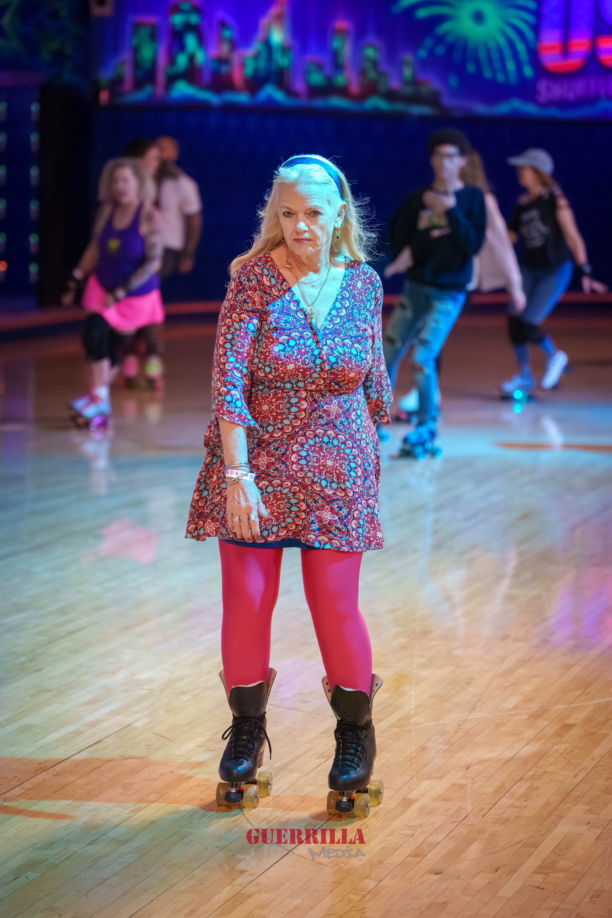 An elderly woman wearing roller skates, a patterned dress, and pink tights is in the foreground of a roller-skating rink with colorful lights. In the background, several younger people are skating and socializing.