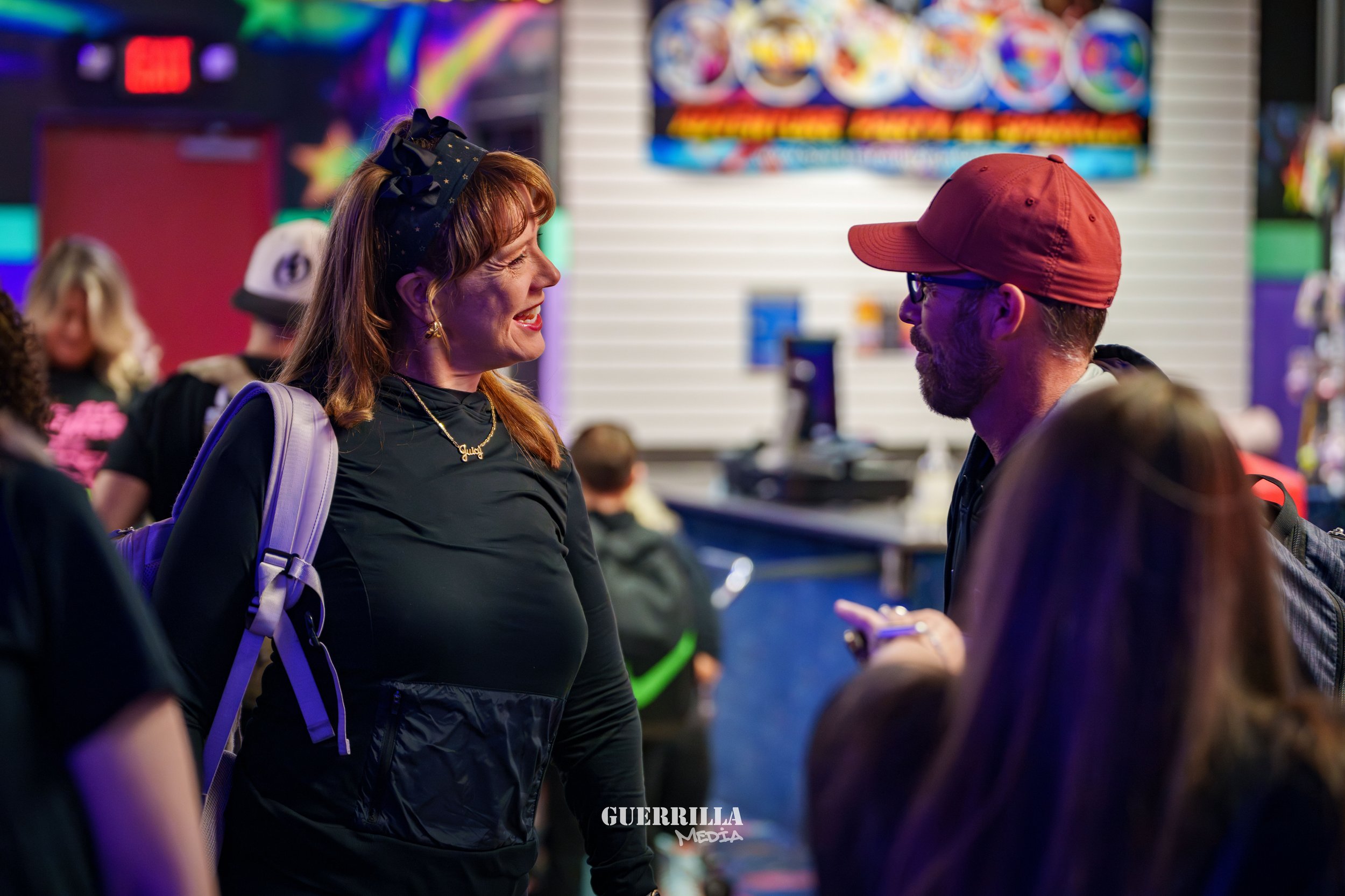 Two people, a woman with red hair wearing a black shirt and a man with a red cap and glasses, are talking at an arcade or entertainment venue. There are other people in the background.