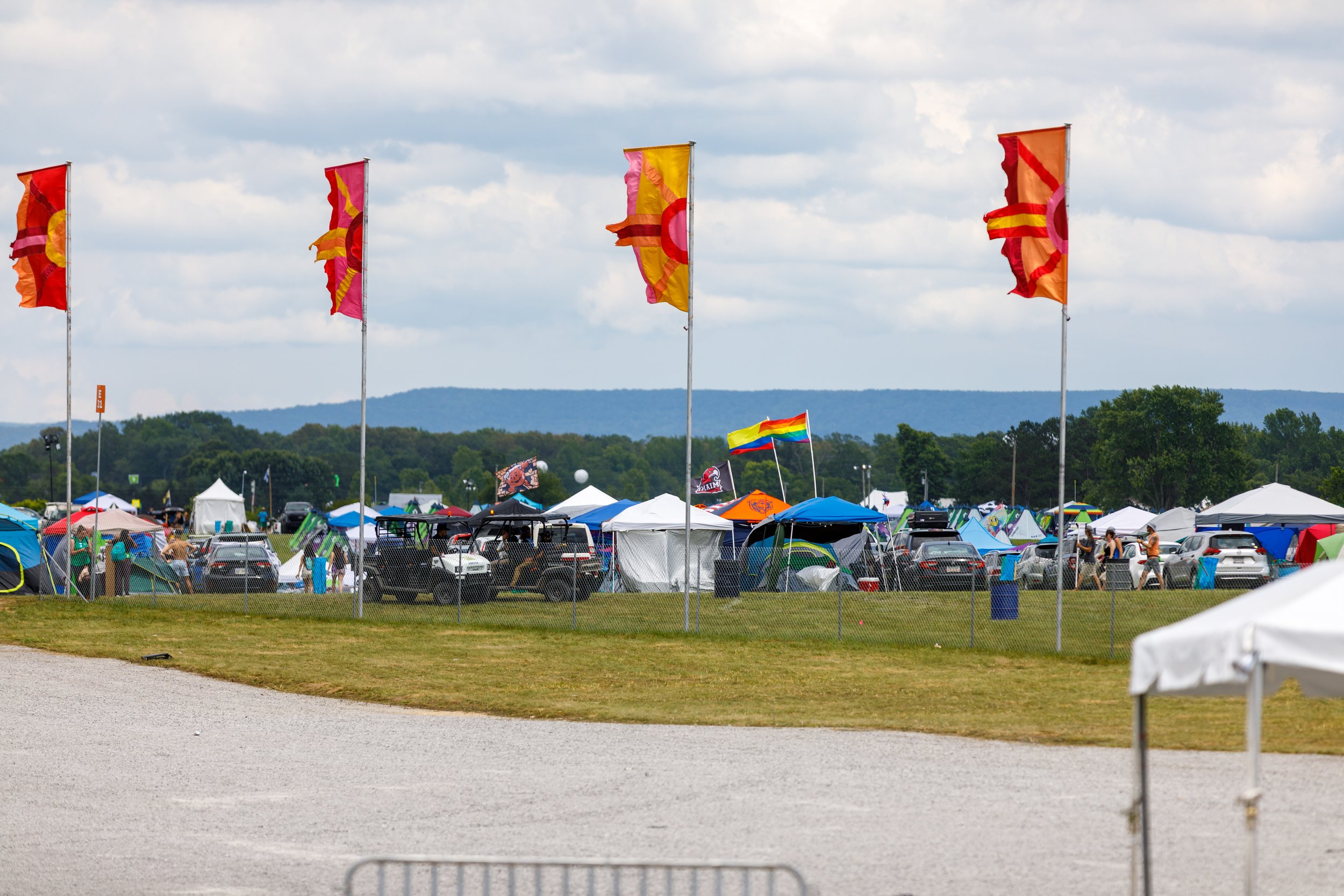 A festival scene with tents, flags, and parked cars on a grassy field under a cloudy sky.