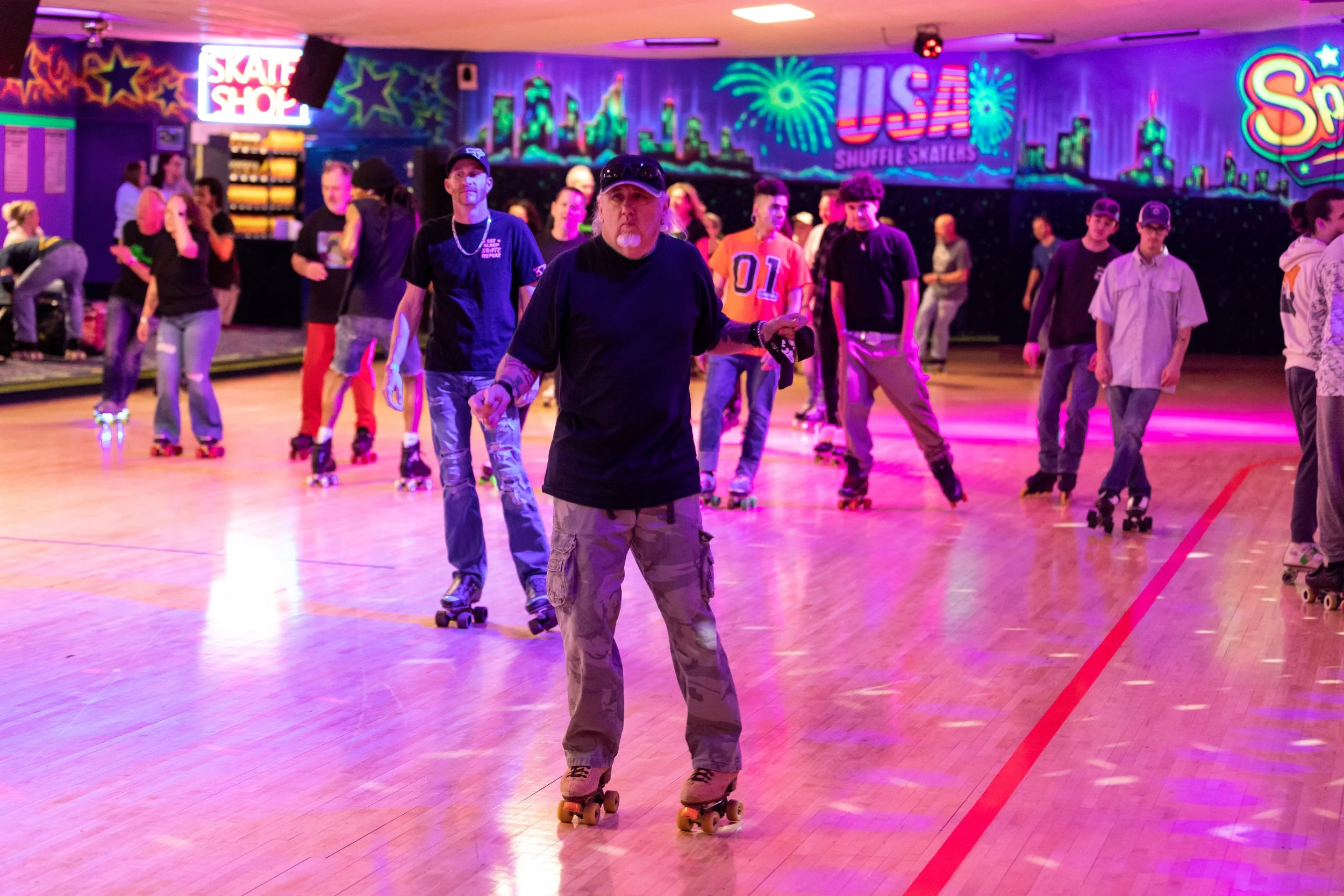 People roller skating inside a neon-lit rink with colorful graffiti on the walls and signs such as 'Skate Shop' and 'USA' with fireworks in the background.