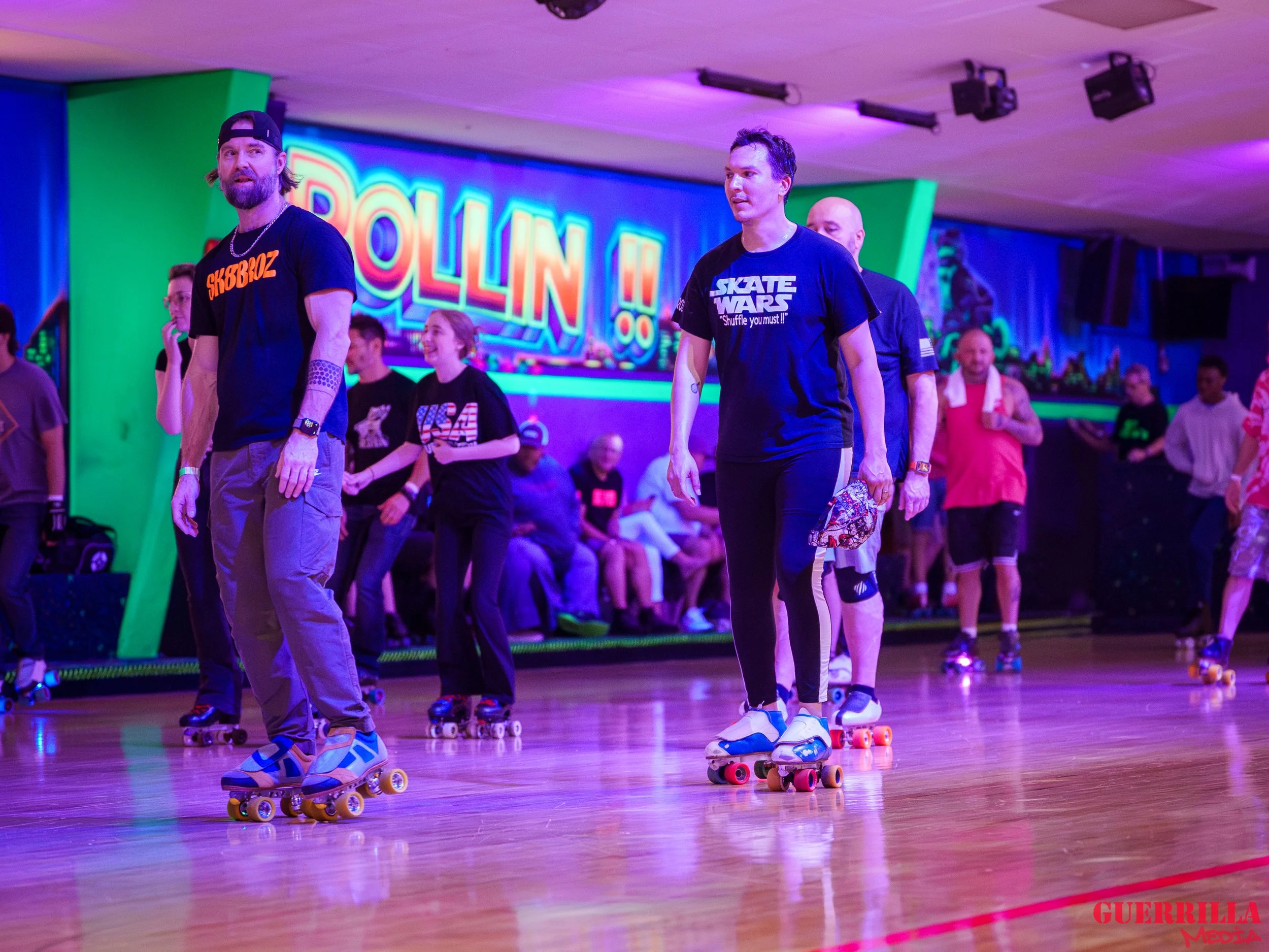 People roller skating at an indoor roller rink with a colorful neon sign in the background that reads "Rolling" and a vibrant mural on the wall.