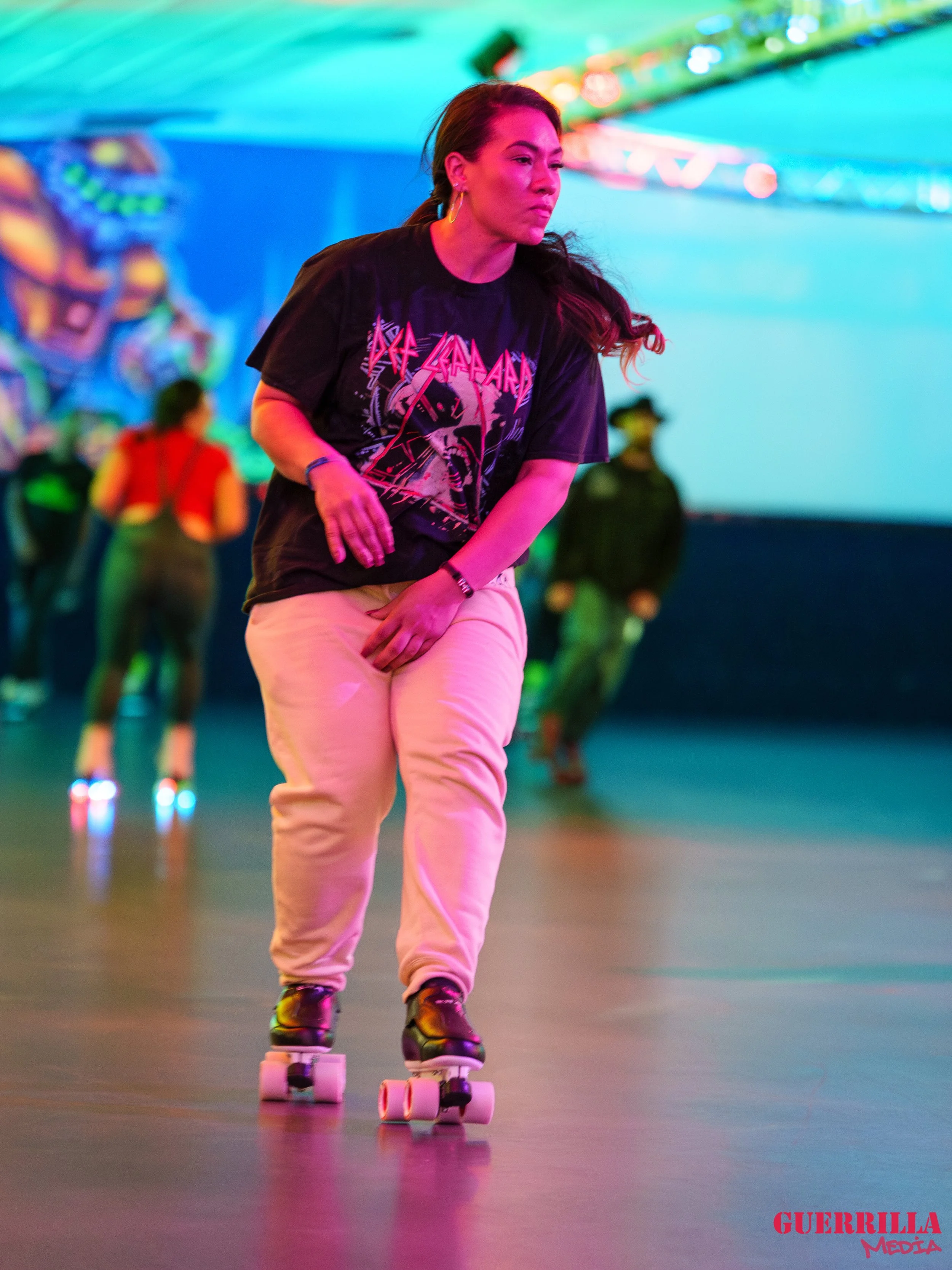 Woman rollerblading indoors near colorful lighting, wearing a black band t-shirt and beige pants.