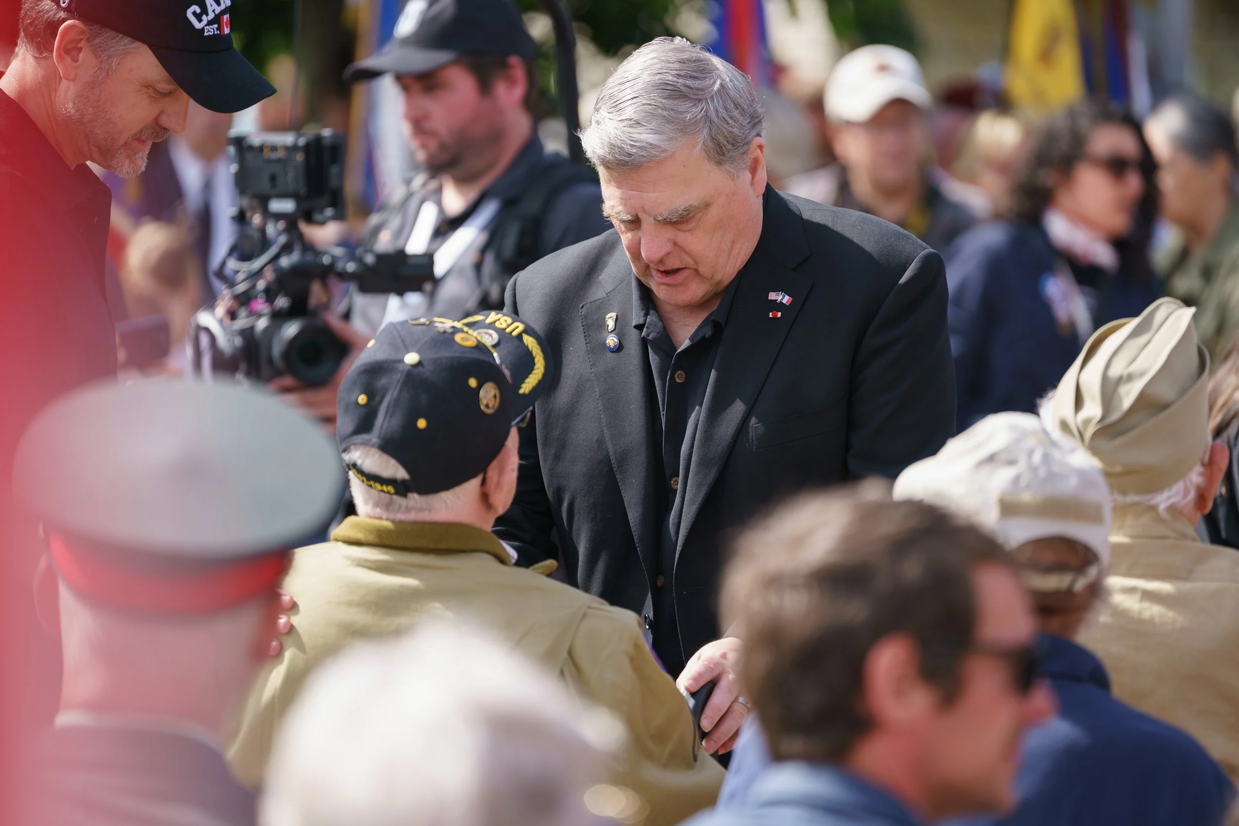 Man in a dark jacket talking to a group of elderly people, some wearing hats, at an outdoor event with many people and greenery in the background.