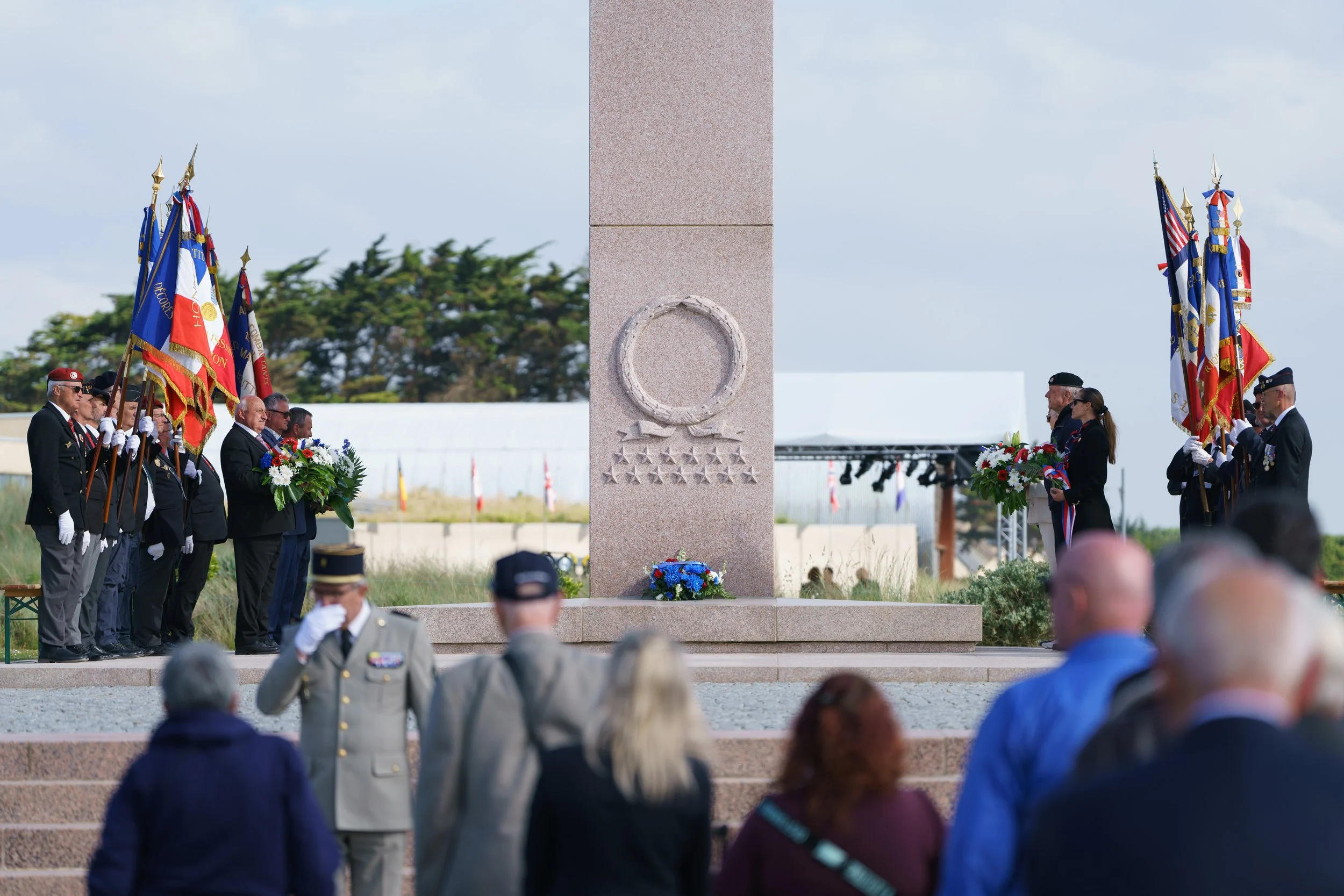 A memorial ceremony with people gathered around a monument, some holding flags and flowers, in an outdoor setting.
