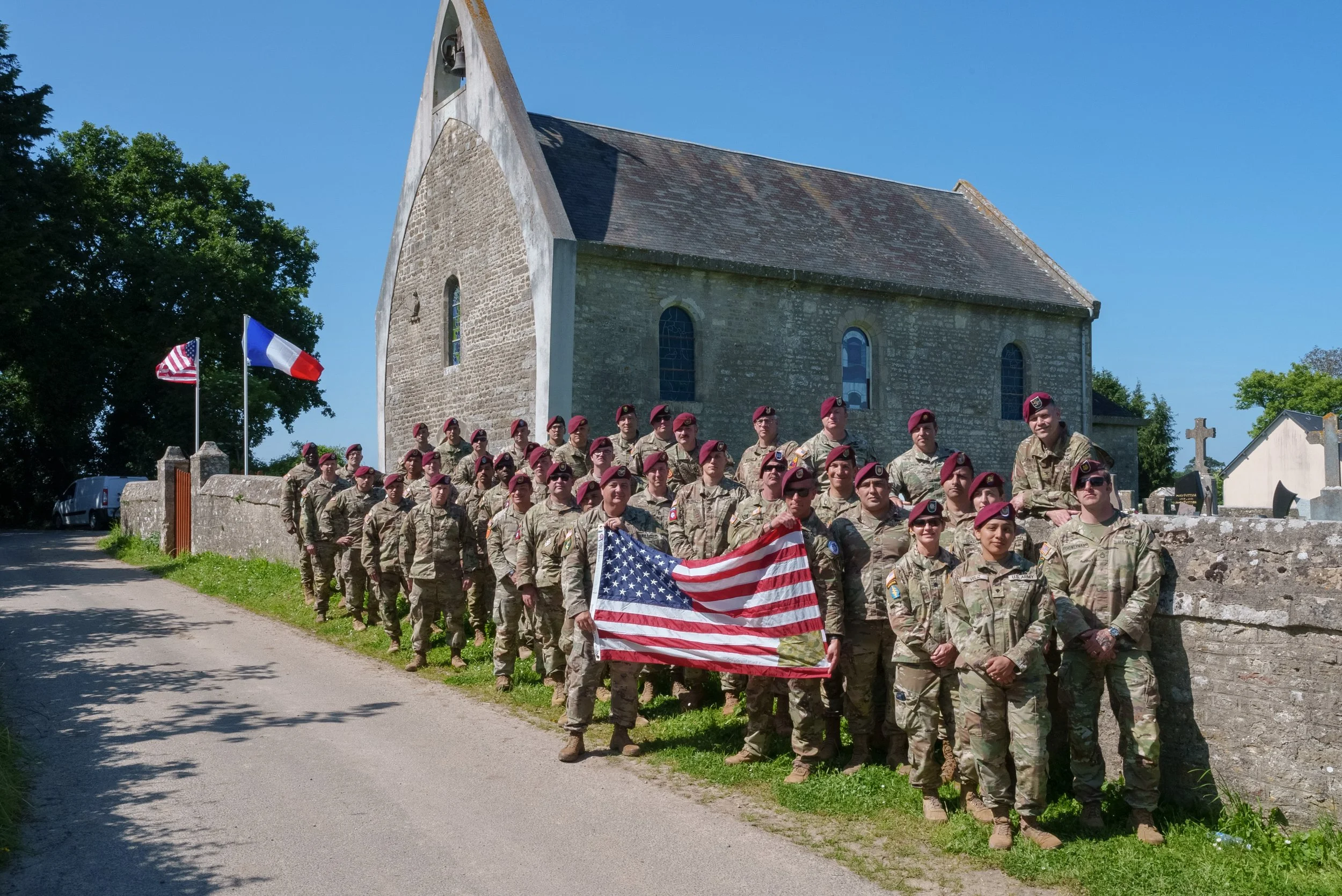Group of soldiers in uniform holding an American flag, standing outside near a stone church building with French flags on a sunny day.