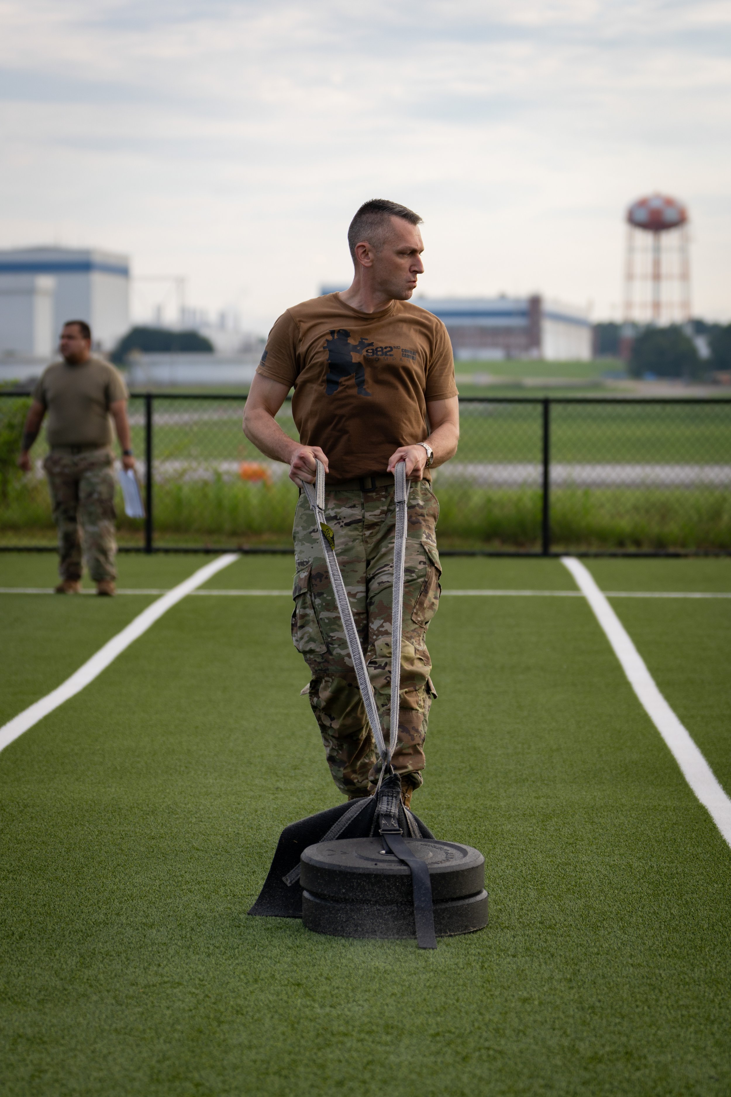 A man in camouflage fatigues and a brown t-shirt is rolling a weighted sled on a green artificial turf field during daytime. Another person in camouflage fatigues is in the background, also on the field, with industrial building structures and a wate