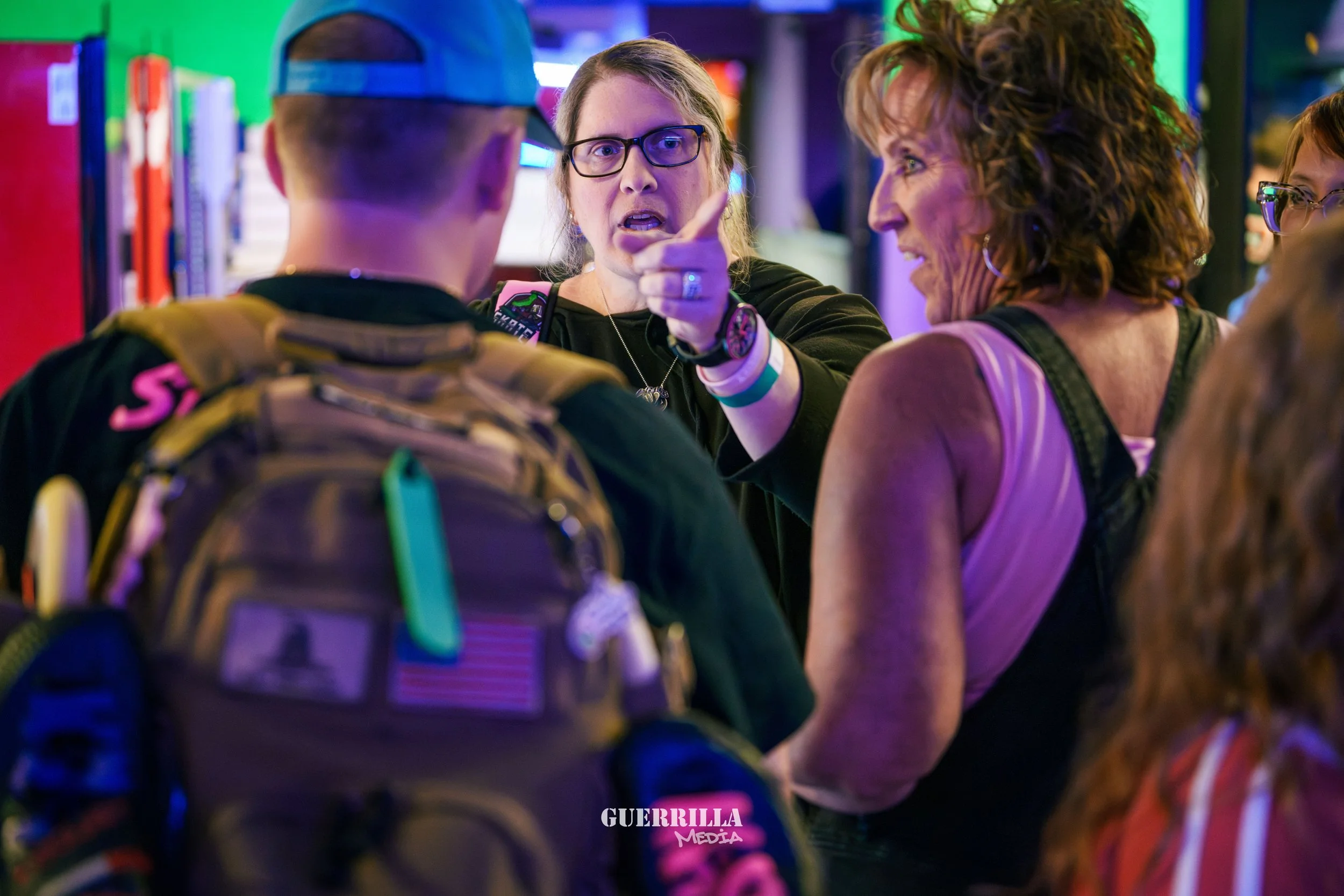 A woman with glasses and long hair is arguing with a young man who is wearing a baseball cap and a backpack. The woman is pointing her finger at the man, and another woman with curly hair and a tank top is standing nearby, observing the interaction. 