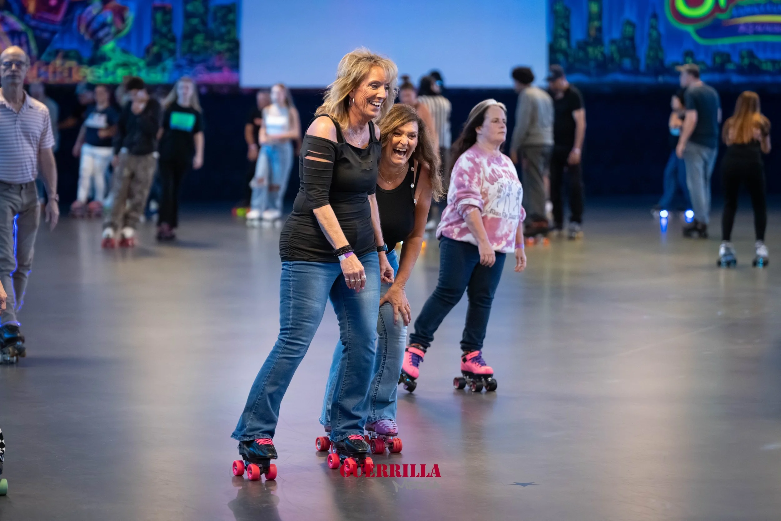 People roller skating in an indoor rink, enjoying and having fun, with some laughing and smiling, colorful mural in the background.