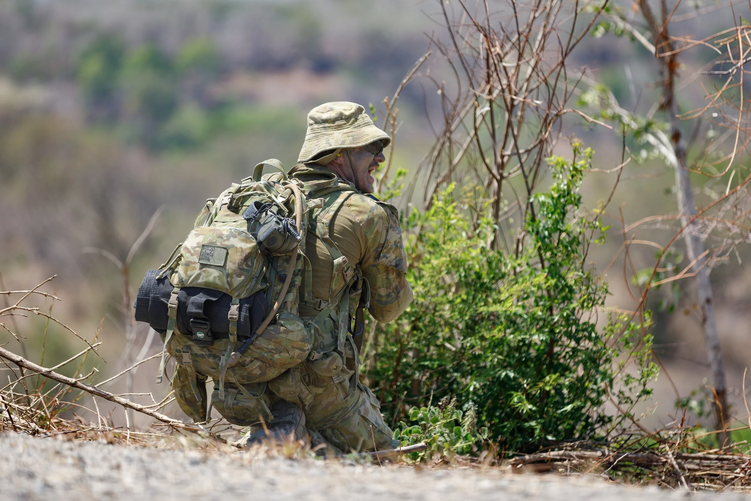 A soldier dressed in camouflage gear, wearing a wide-brimmed hat, kneeling next to a bush in a dry, open landscape.