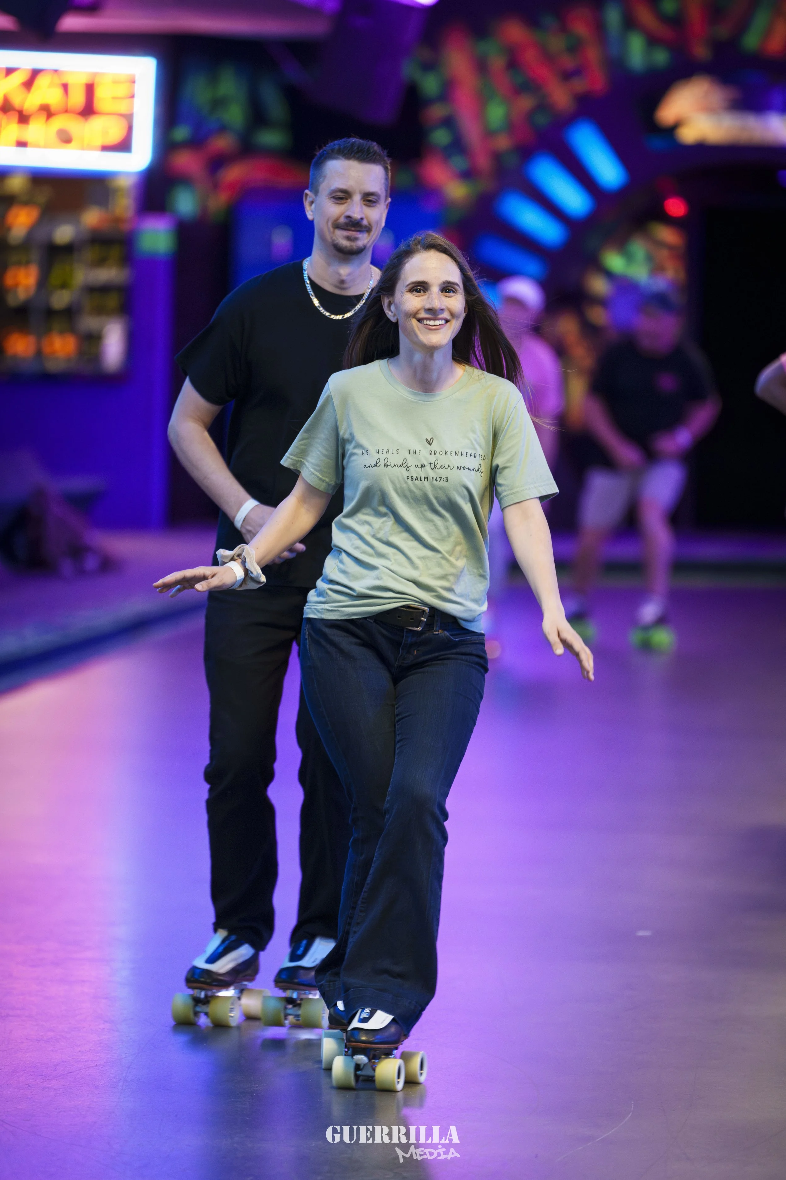 A young woman roller skating indoors with a man behind her, smiling and balancing, with colorful neon lights and people in the background.