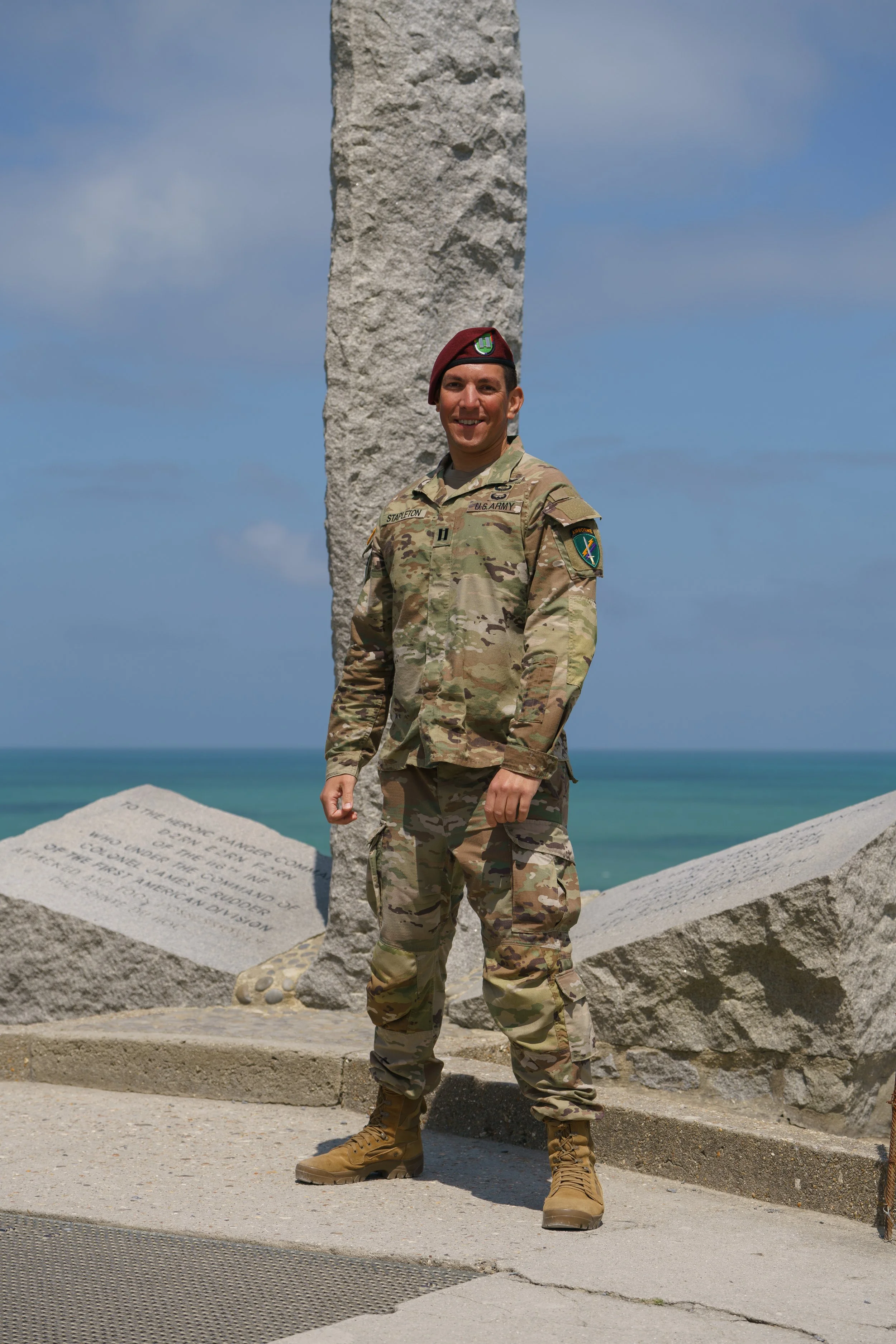 A soldier in camouflage uniform and combat boots standing outdoors in front of a large stone monument with plaques, near a beach with the ocean and a blue sky in the background.