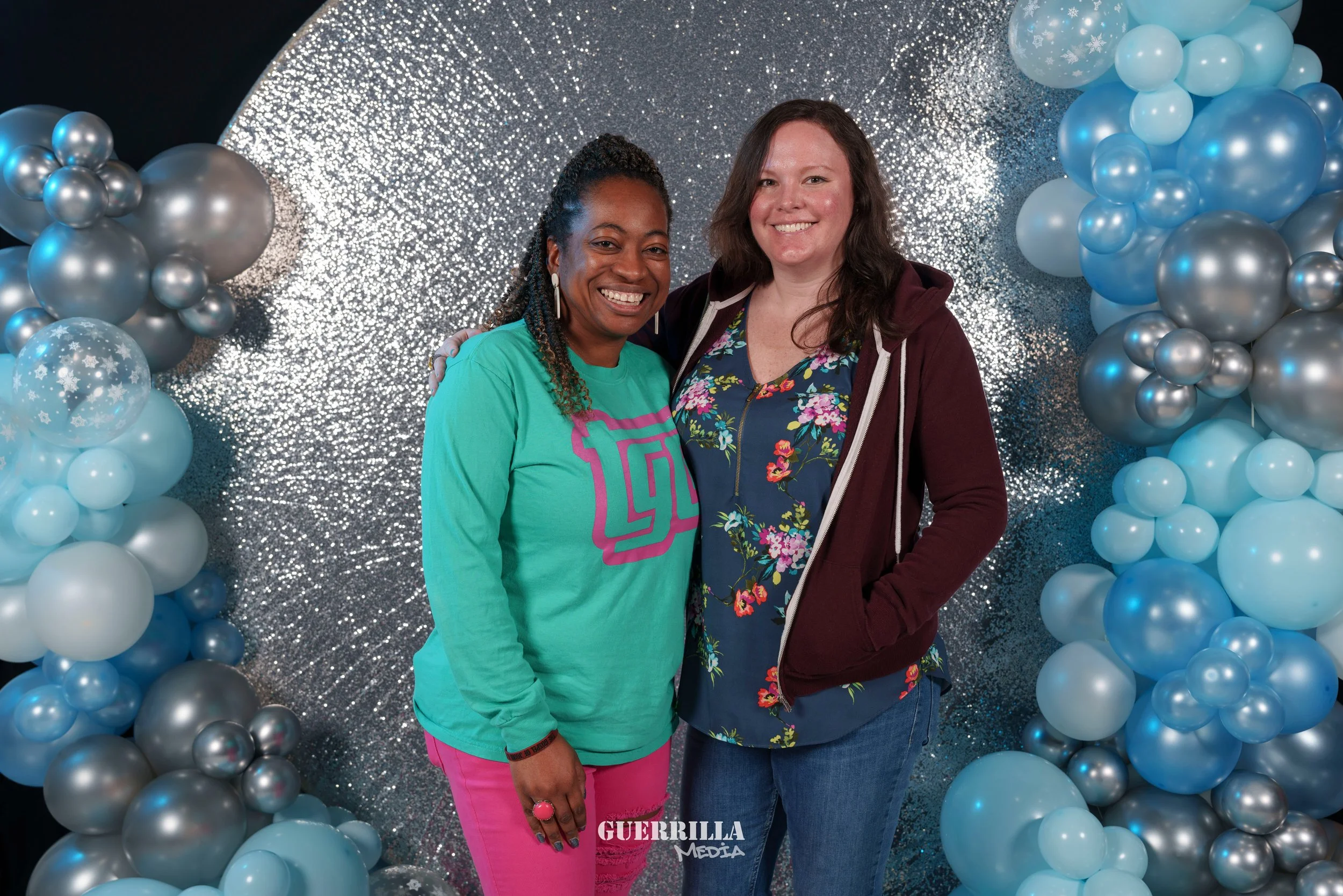 Two women smiling and standing together in front of a silver glitter backdrop with balloon decorations, one wears a green shirt with pink pants, the other wears a floral blouse with a maroon jacket.