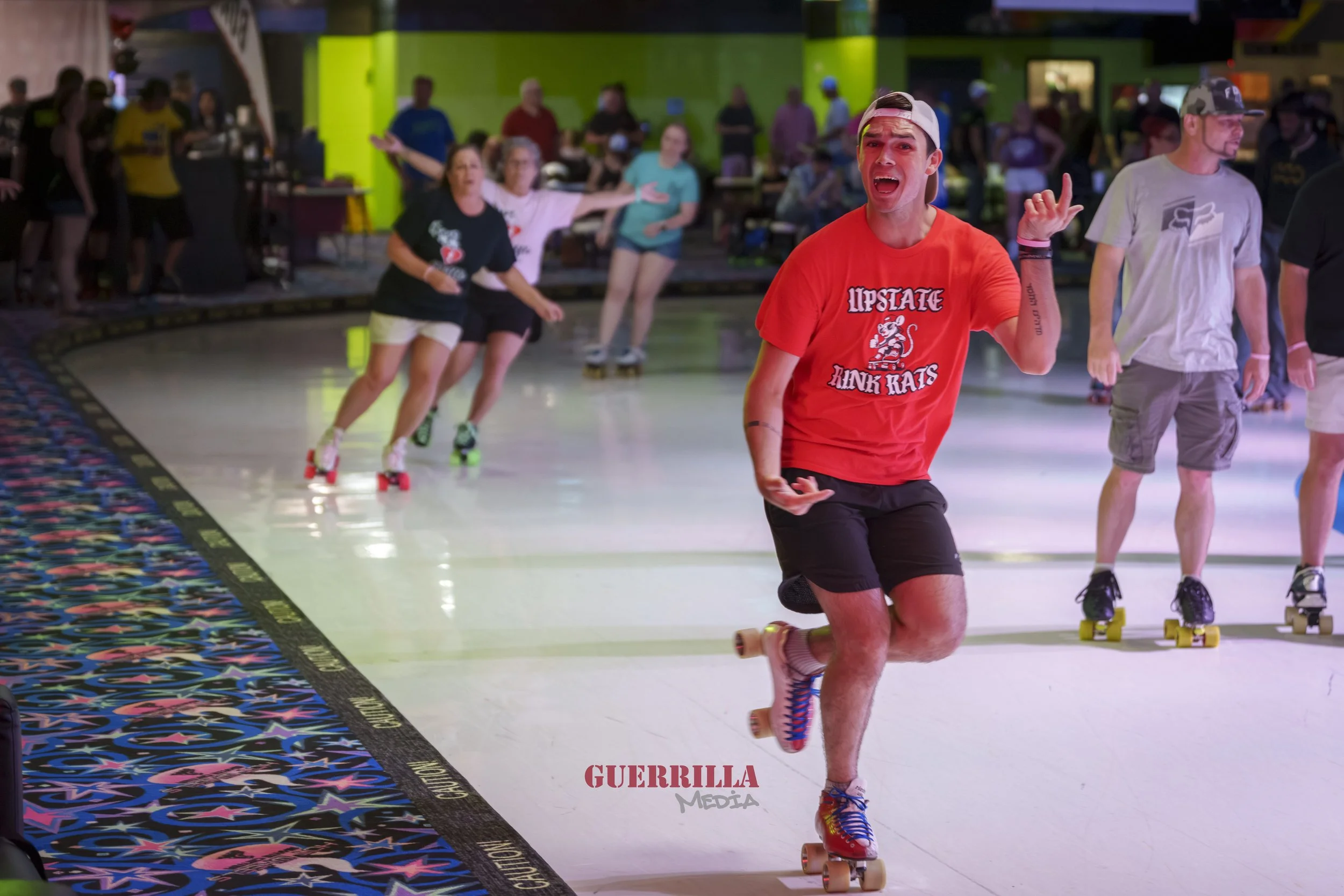 A young man roller skating indoors, wearing a red t-shirt that says 'Hip State Think Rats,' with other people skateboarding and socializing in the background.