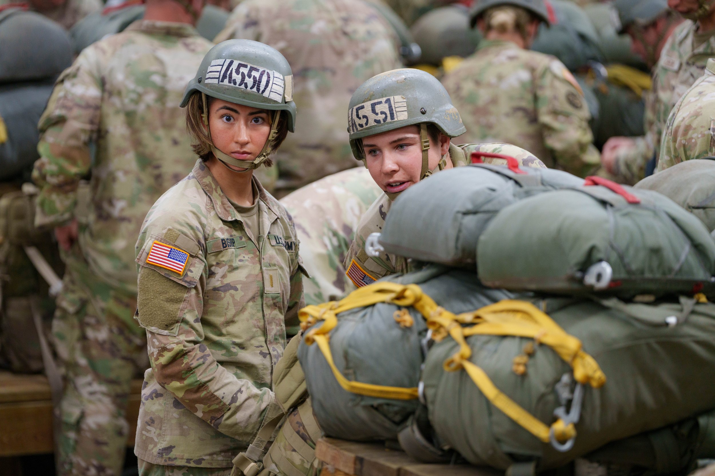 Two young female soldiers in US military uniform and helmets surrounded by fellow soldiers, handling large packs and gear.