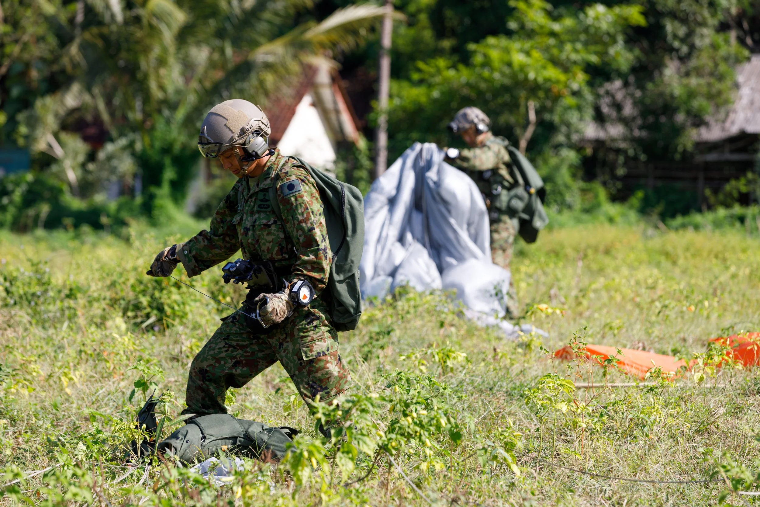 Two soldiers in camouflage uniforms and helmets walking through a grassy field with trees and houses in the background.