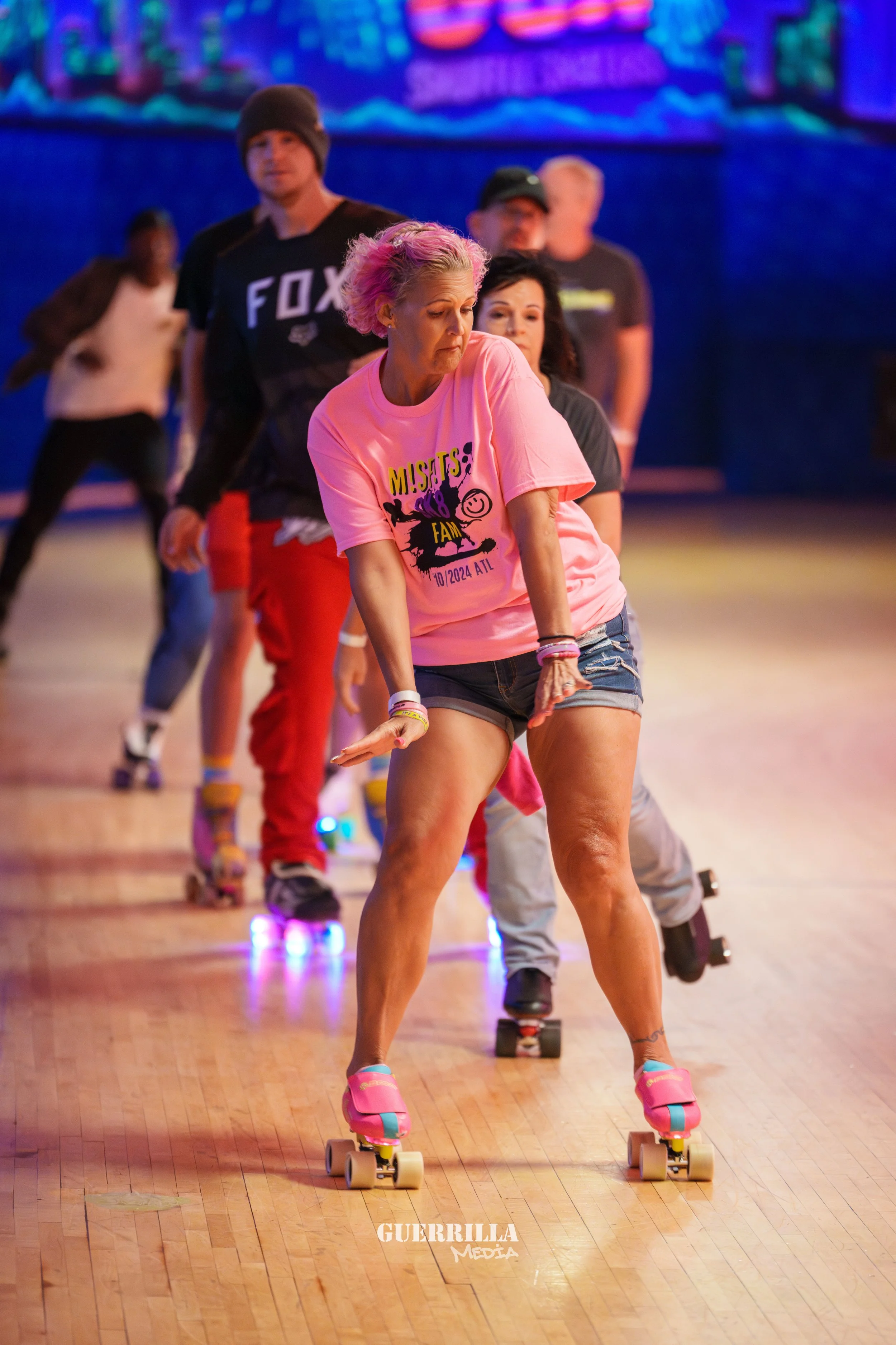 Group of people roller skating indoors, with a woman in the foreground wearing a pink t-shirt and denim shorts.