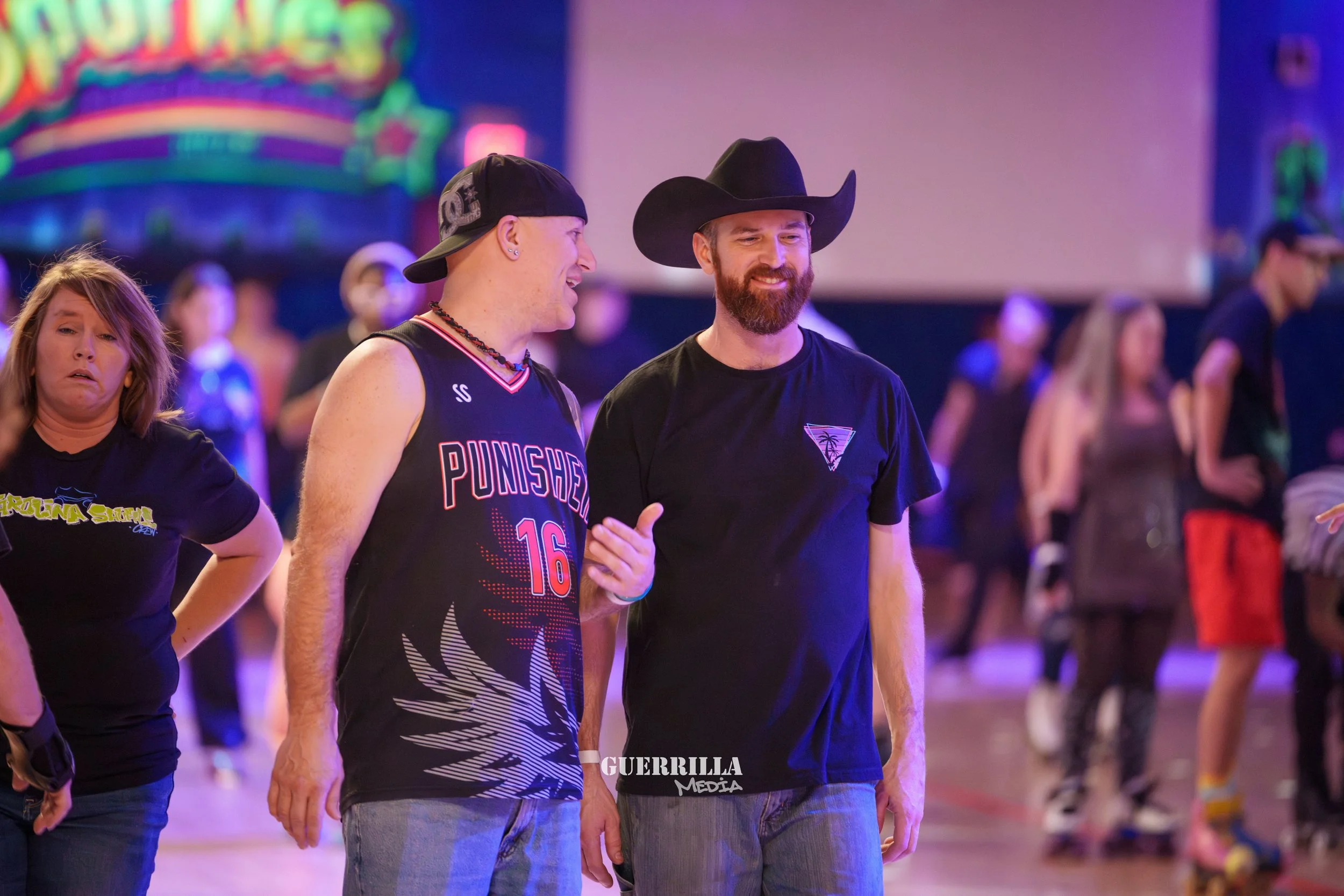 Two men are skating at an indoor roller rink, with one wearing a black basketball jersey and the other wearing a black t-shirt with a triangle logo and a cowboy hat. Several other skaters are in the background.