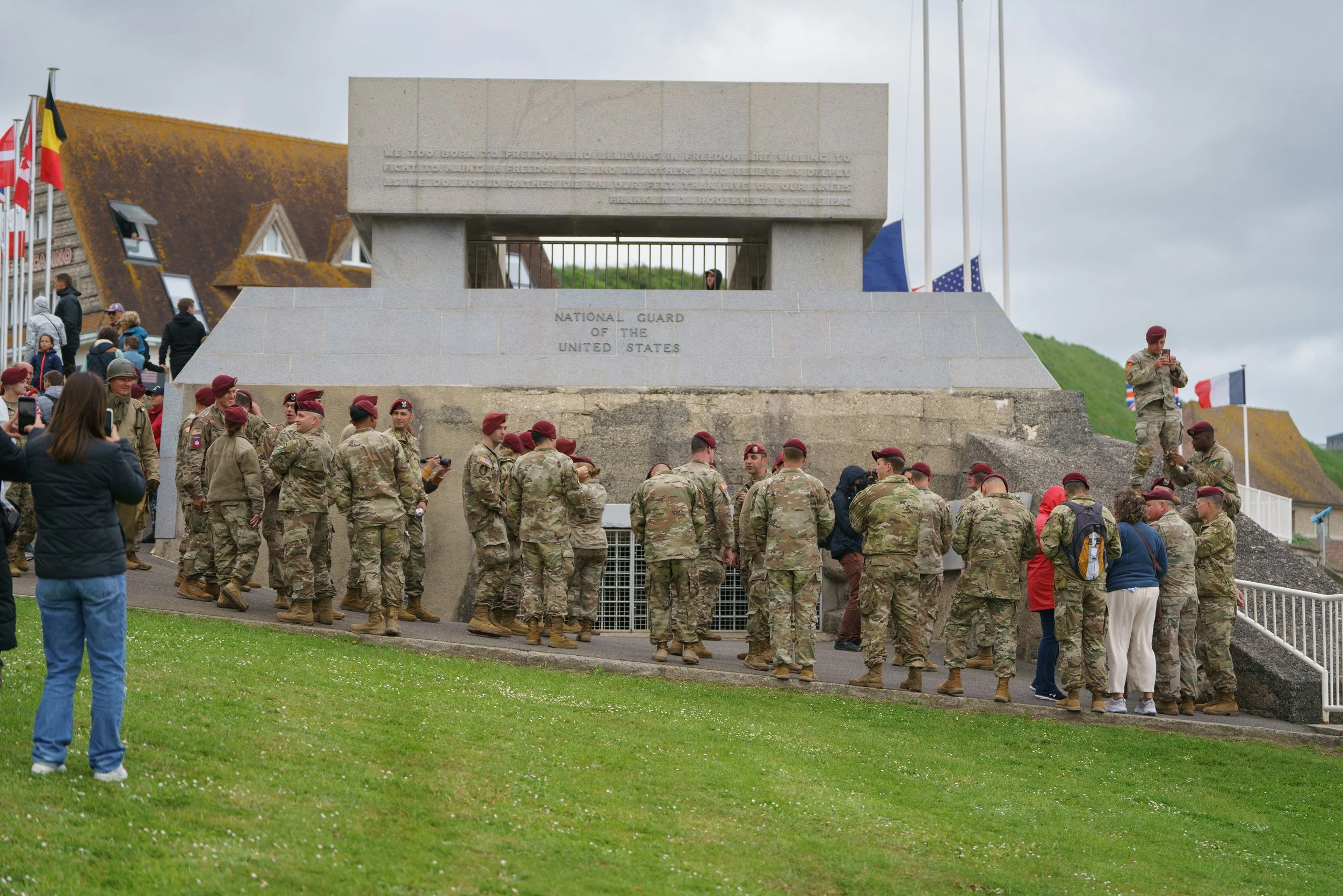 Group of soldiers in uniform with red berets gathered around the Normandy American Cemetery and Memorial. Some are taking photos, and others are looking at the memorial, which features a large stone structure with inscriptions, surrounded by flags an
