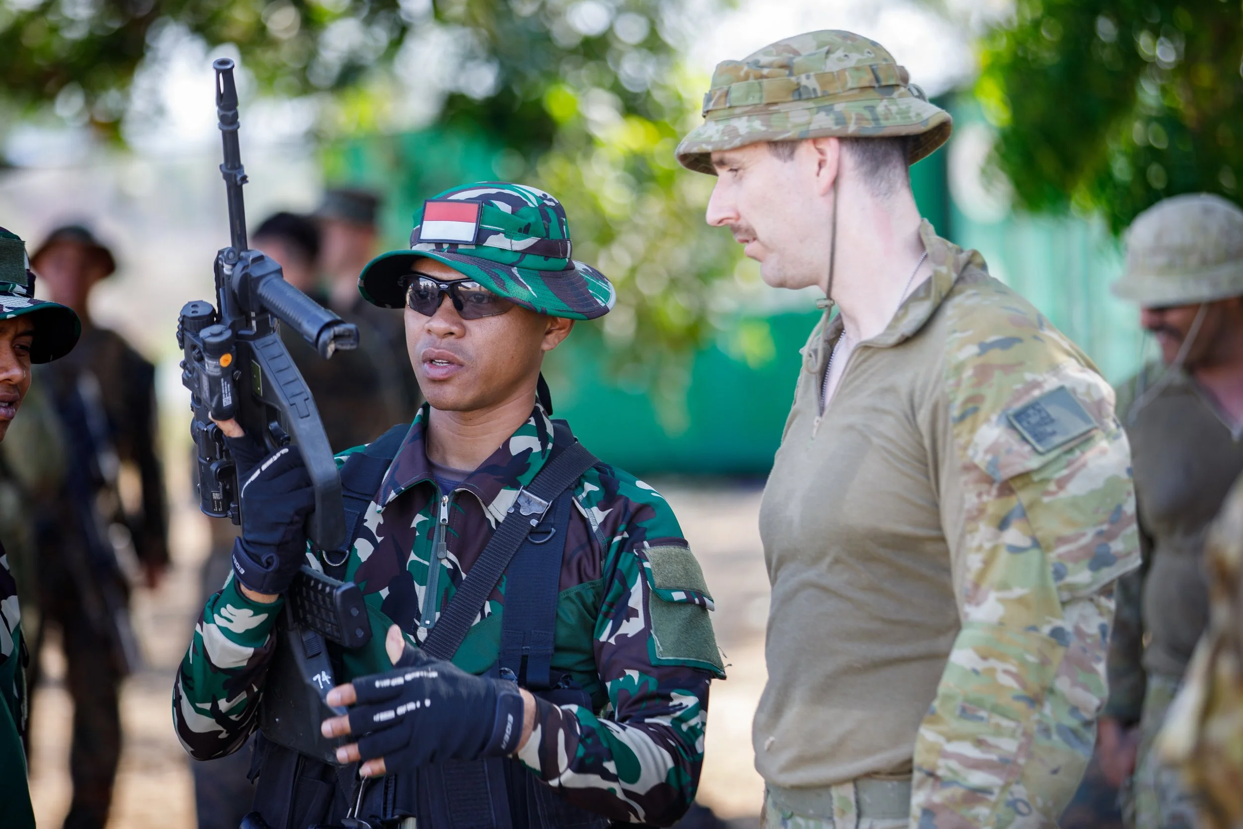 Military personnel in uniform, some wearing camouflage and one in a tan uniform, engaged in a discussion outdoors during the daytime, with trees in the background.